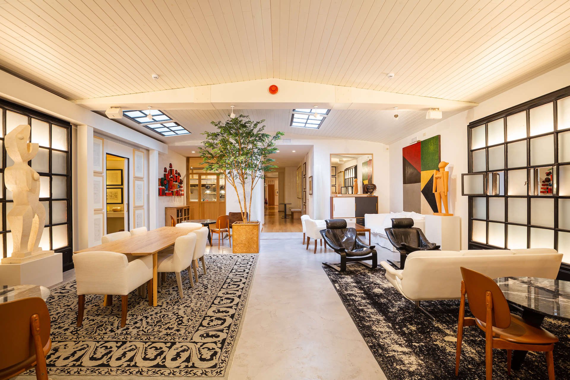 The lounge at Margot House, a long white room with a skylight, a tree in a planter, armchairs, a long table and modern art on the walls