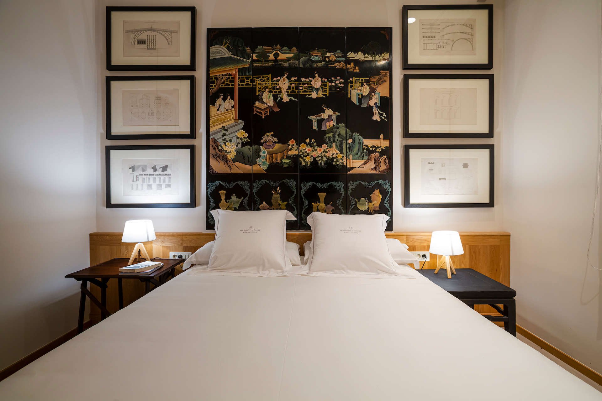Guest room at Margot House with a black lacquered screen behind the bed, framed architectural drawings and white bedding