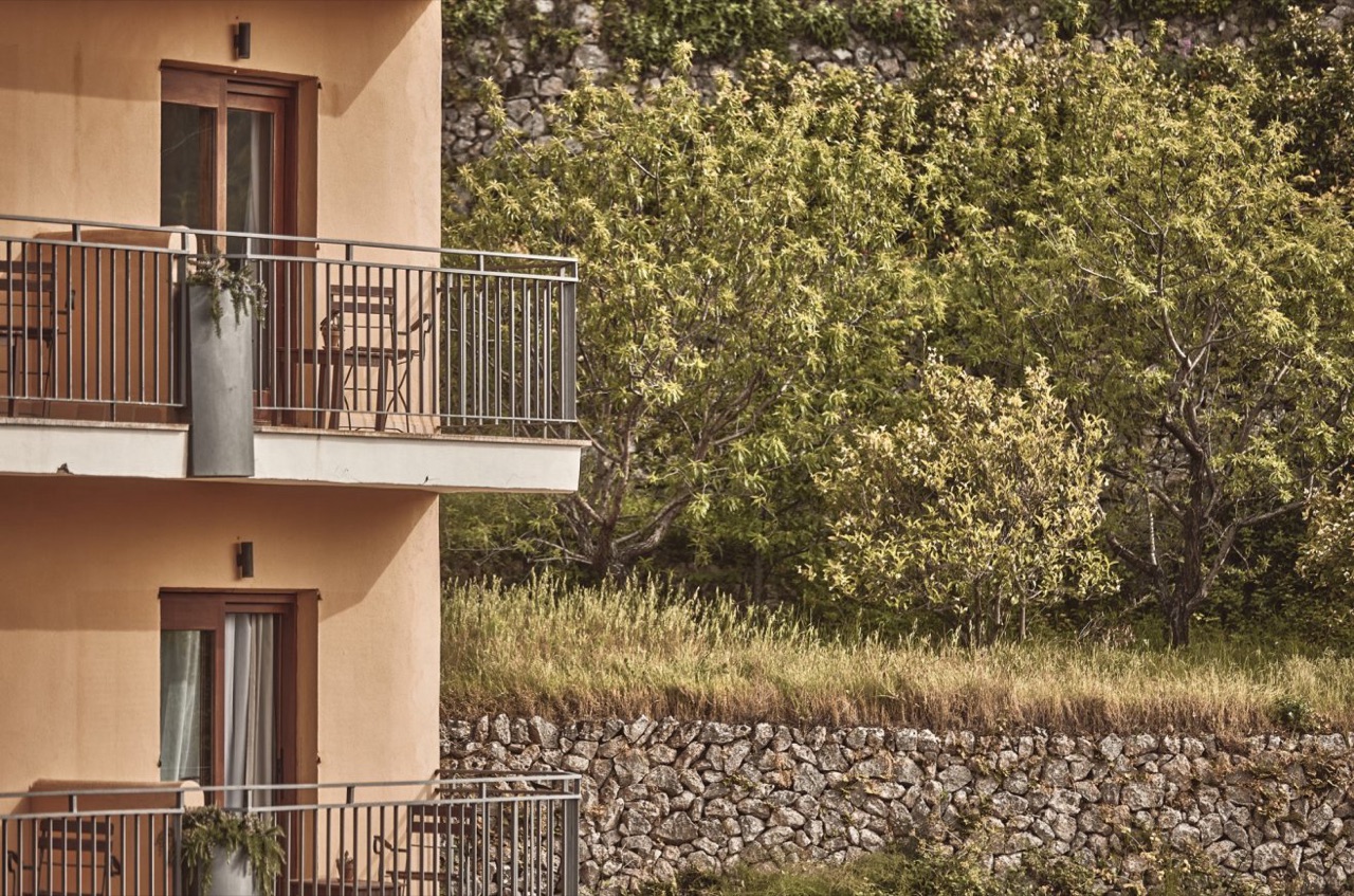 Terracotta-colored hotel facade with private balconies, set against a stone-terraced hillside planted with trees.