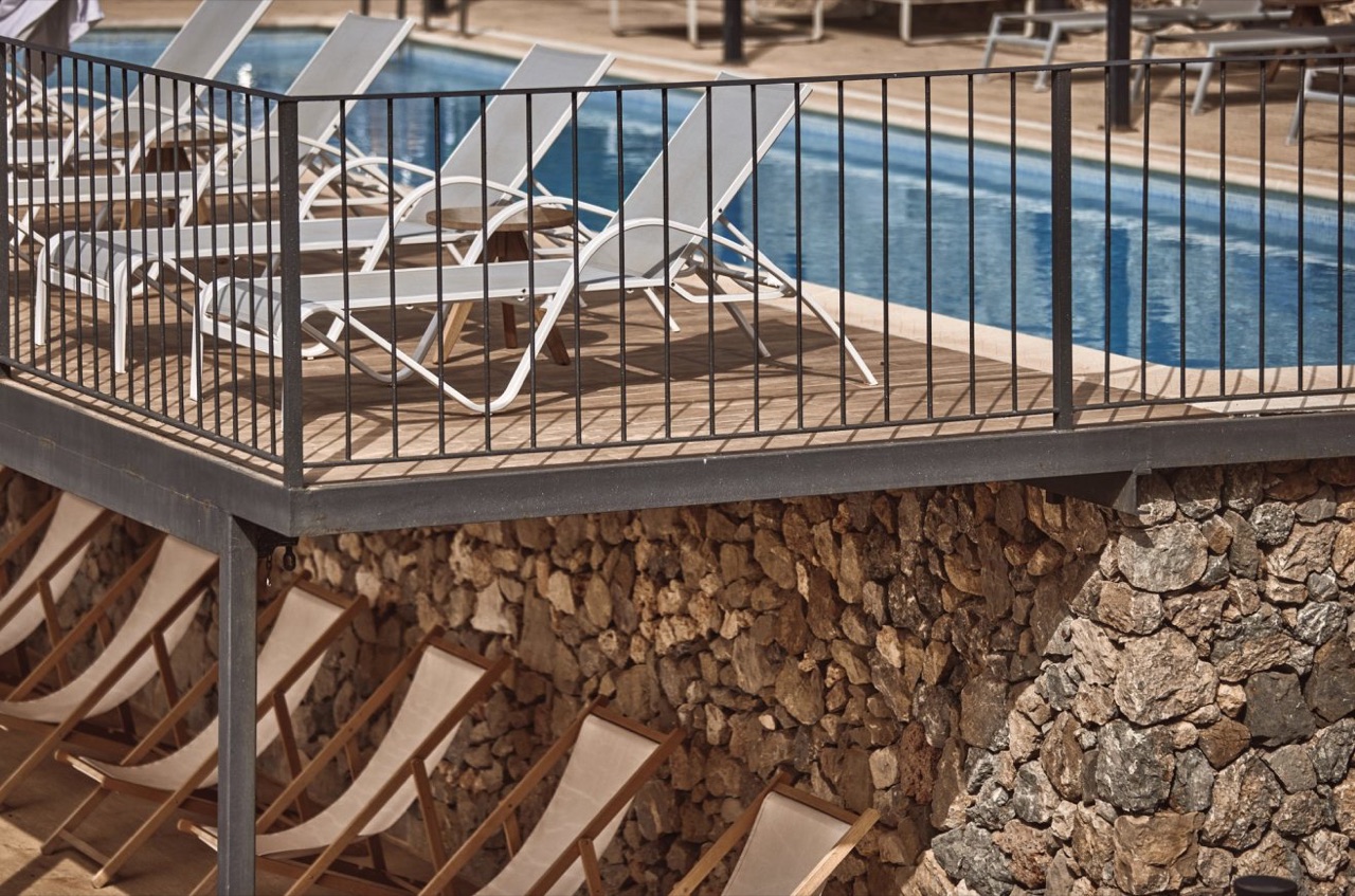 Outdoor swimming pool deck with sun loungers, seen above a stone retaining wall.