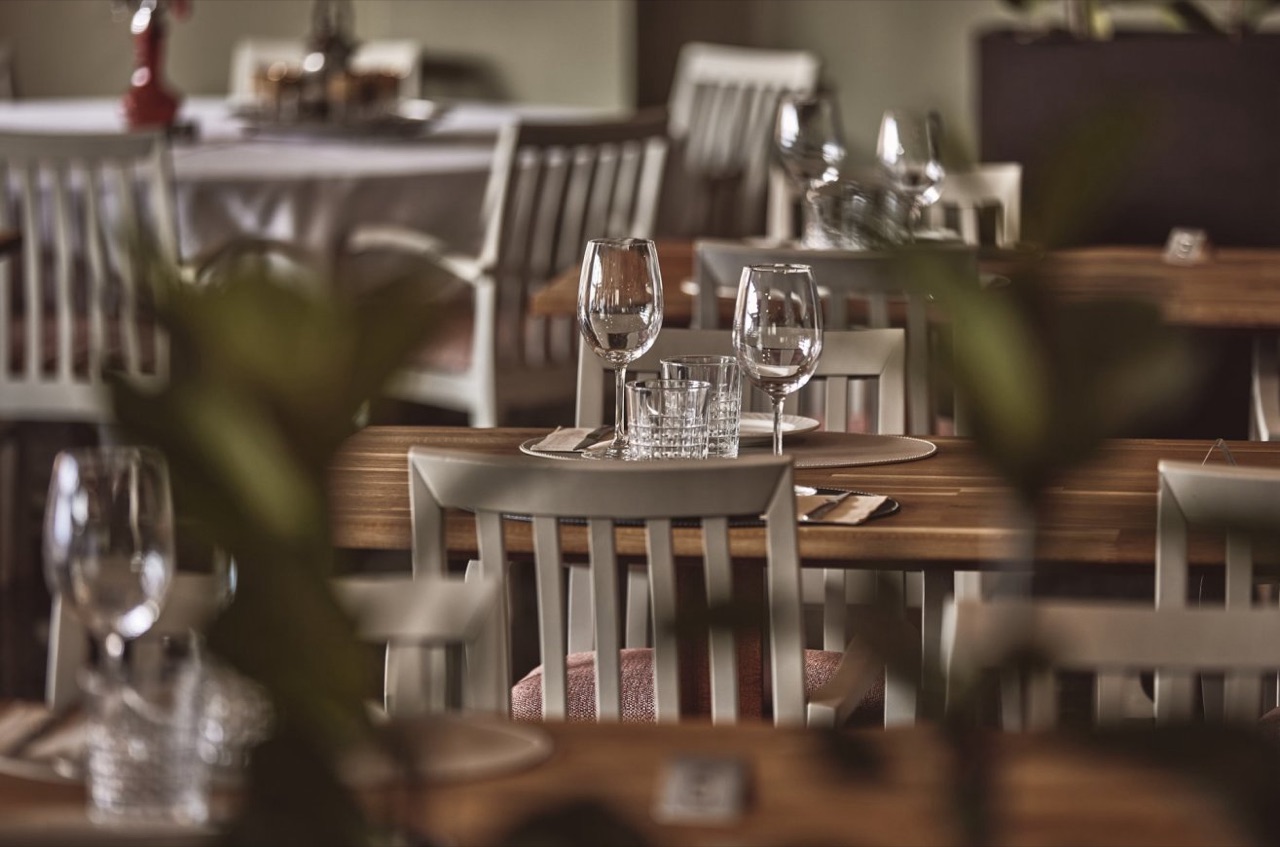 Bistro dining room with wine glasses set on wooden tables and pale grey chairs, seen through potted plants.