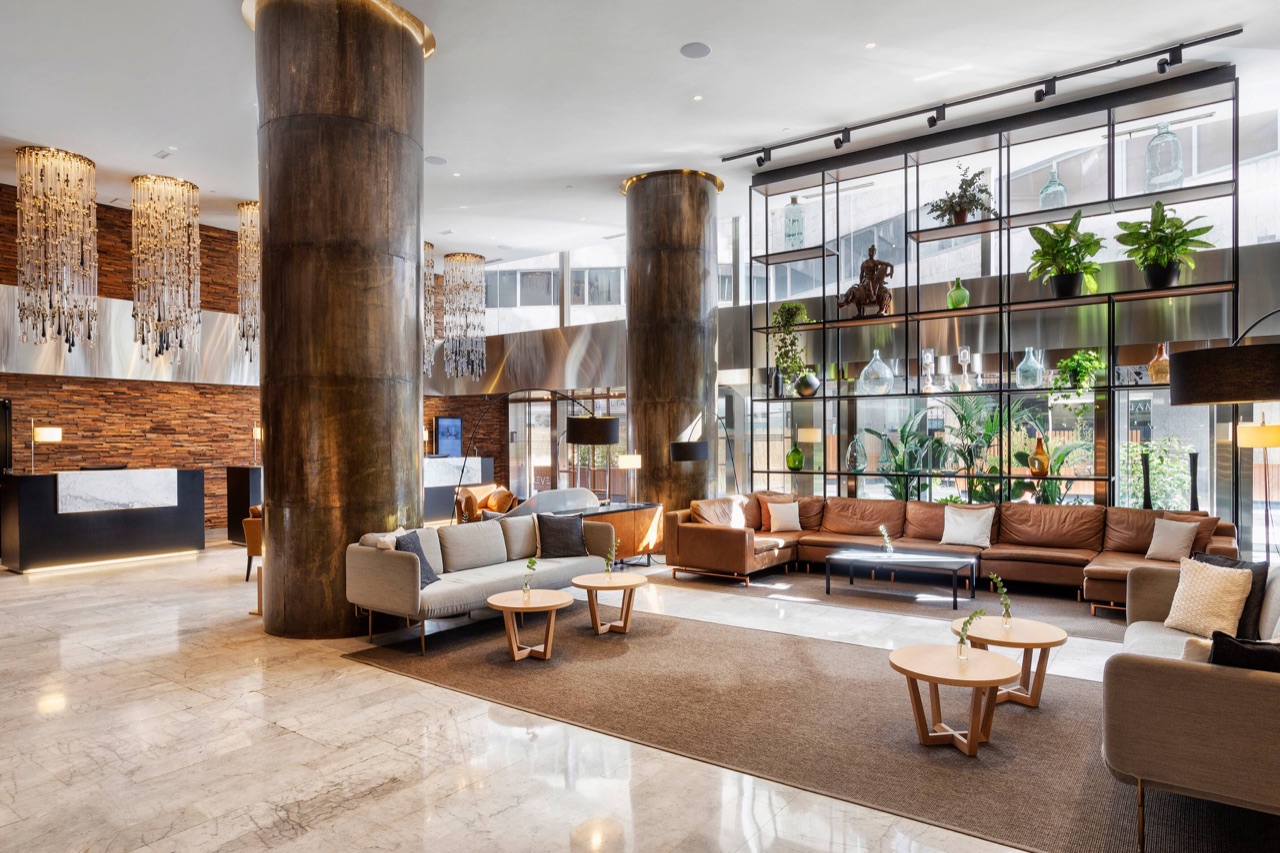 Meliá Castilla's lobby with large metal columns, hanging crystal chandeliers, leather sofas and a glass display shelf of bottles and plants.