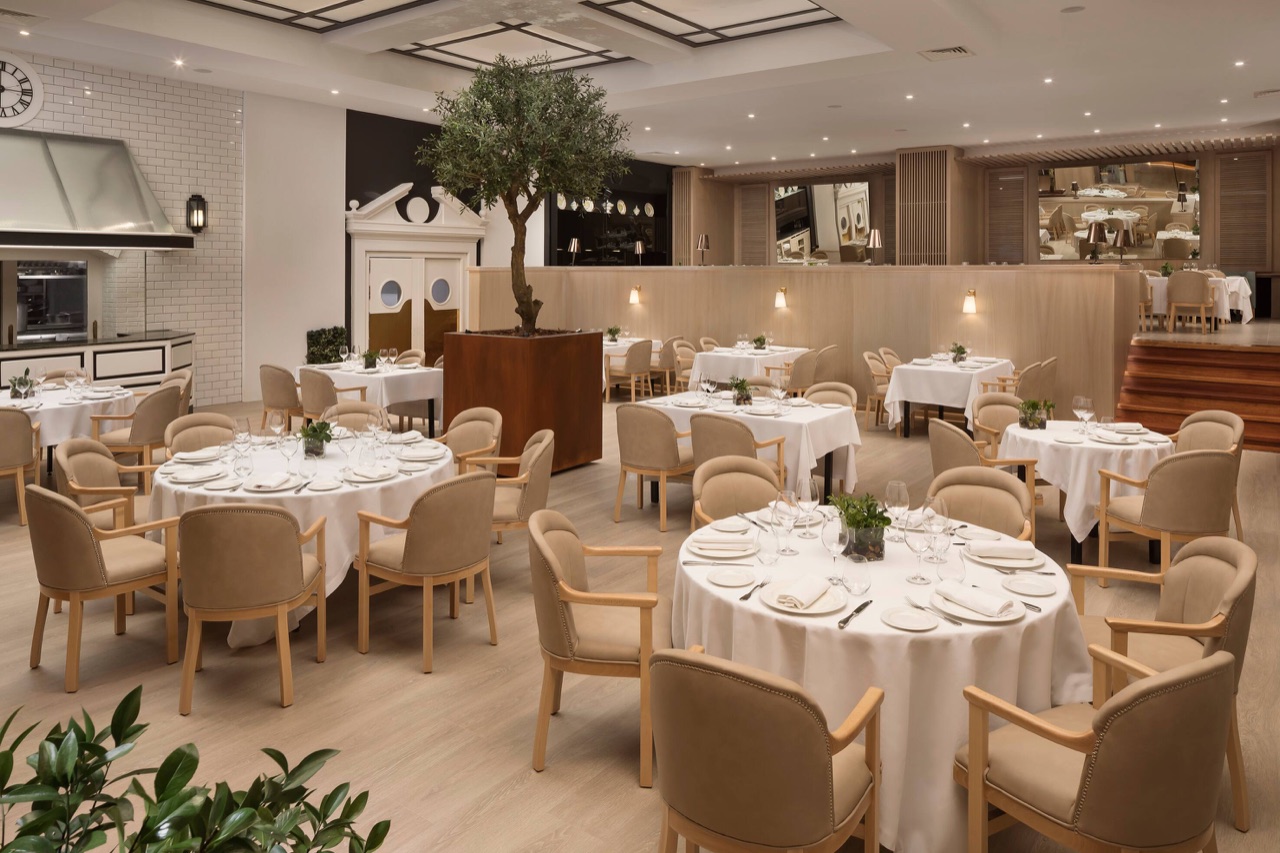 The L'Albufera dining room at Meliá Castilla, with round tables set in white linen, beige upholstered chairs and a potted olive tree at the centre.