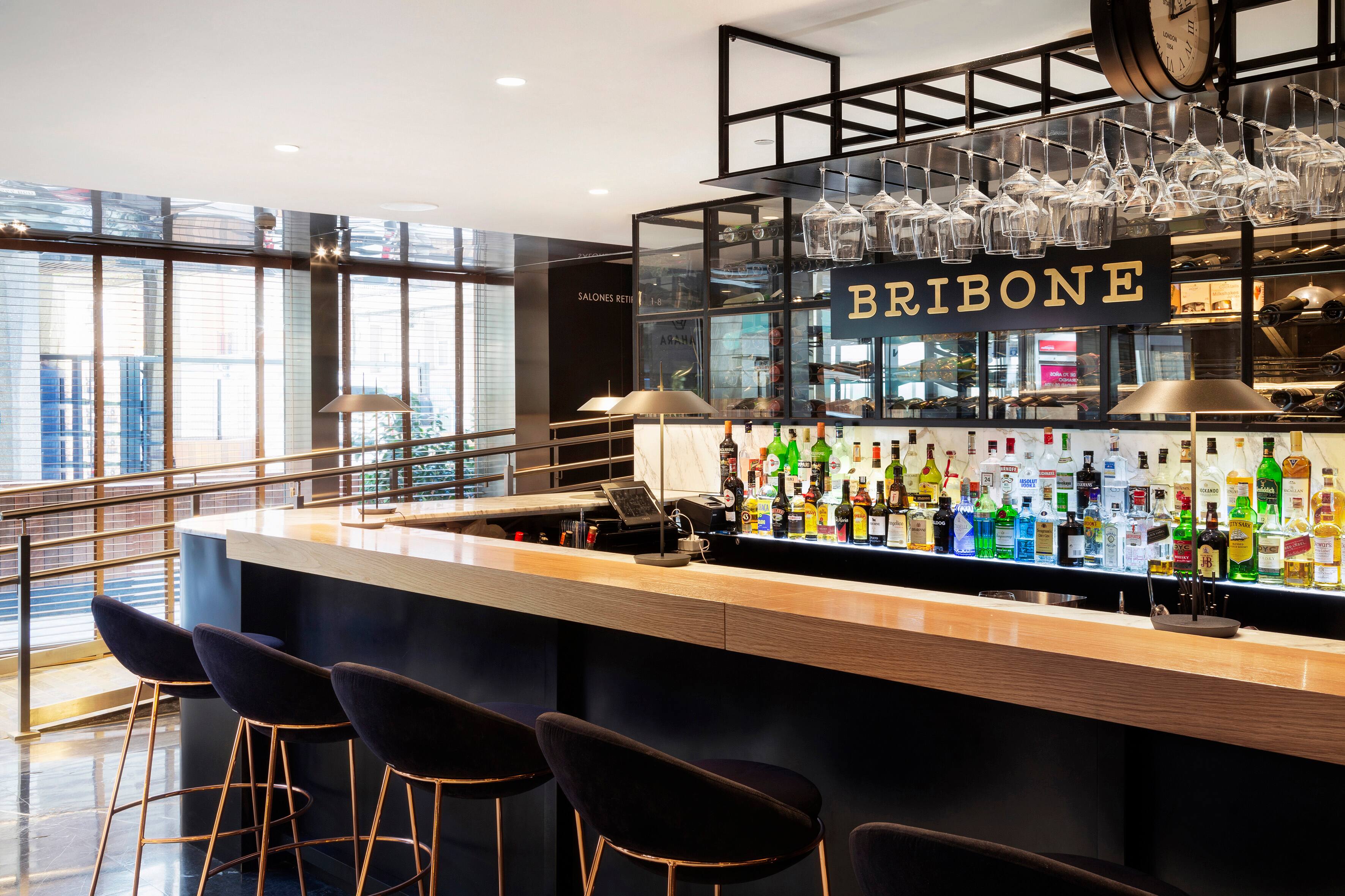 A bar counter in pale wood with dark stools, wine glasses hanging above and a backlit shelf of spirits bottles beneath a sign reading BRIBONE.