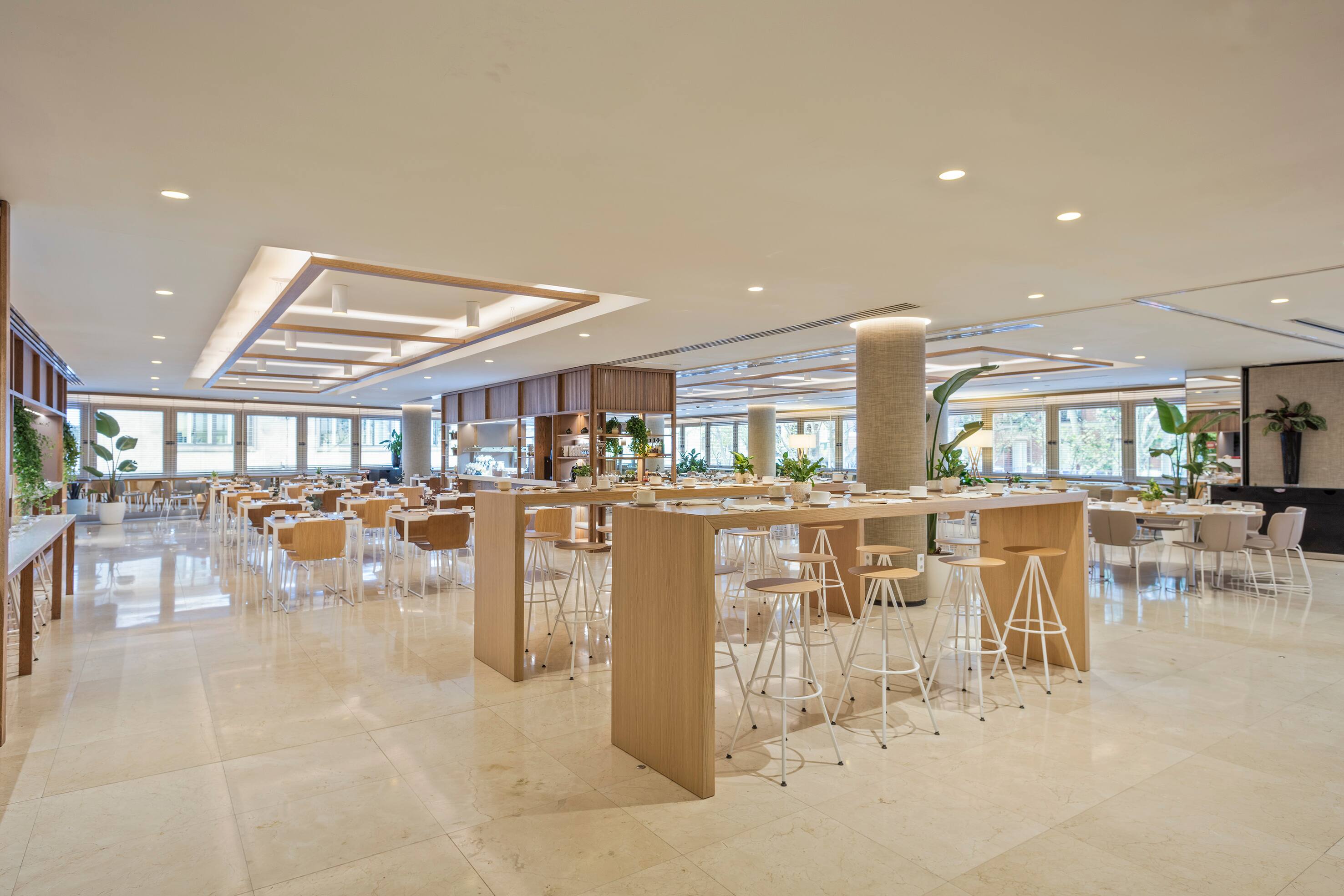 A bright dining room with pale marble floors, rows of set tables, a tall shared wood counter with stools and windows along two walls.