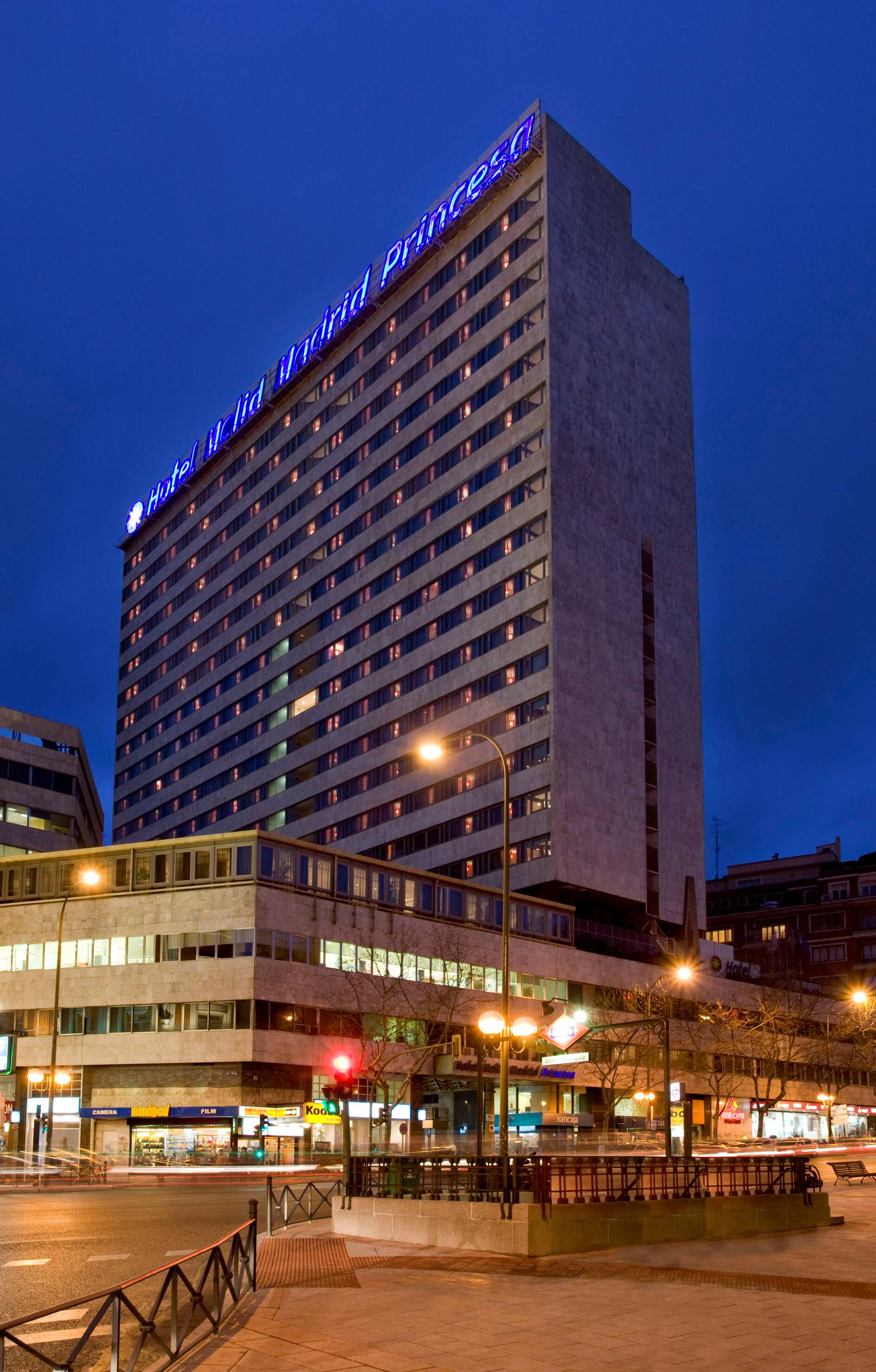 A tall hotel tower photographed at dusk with an illuminated blue rooftop sign reading Hotel Melia Madrid Princesa above a lit street corner.