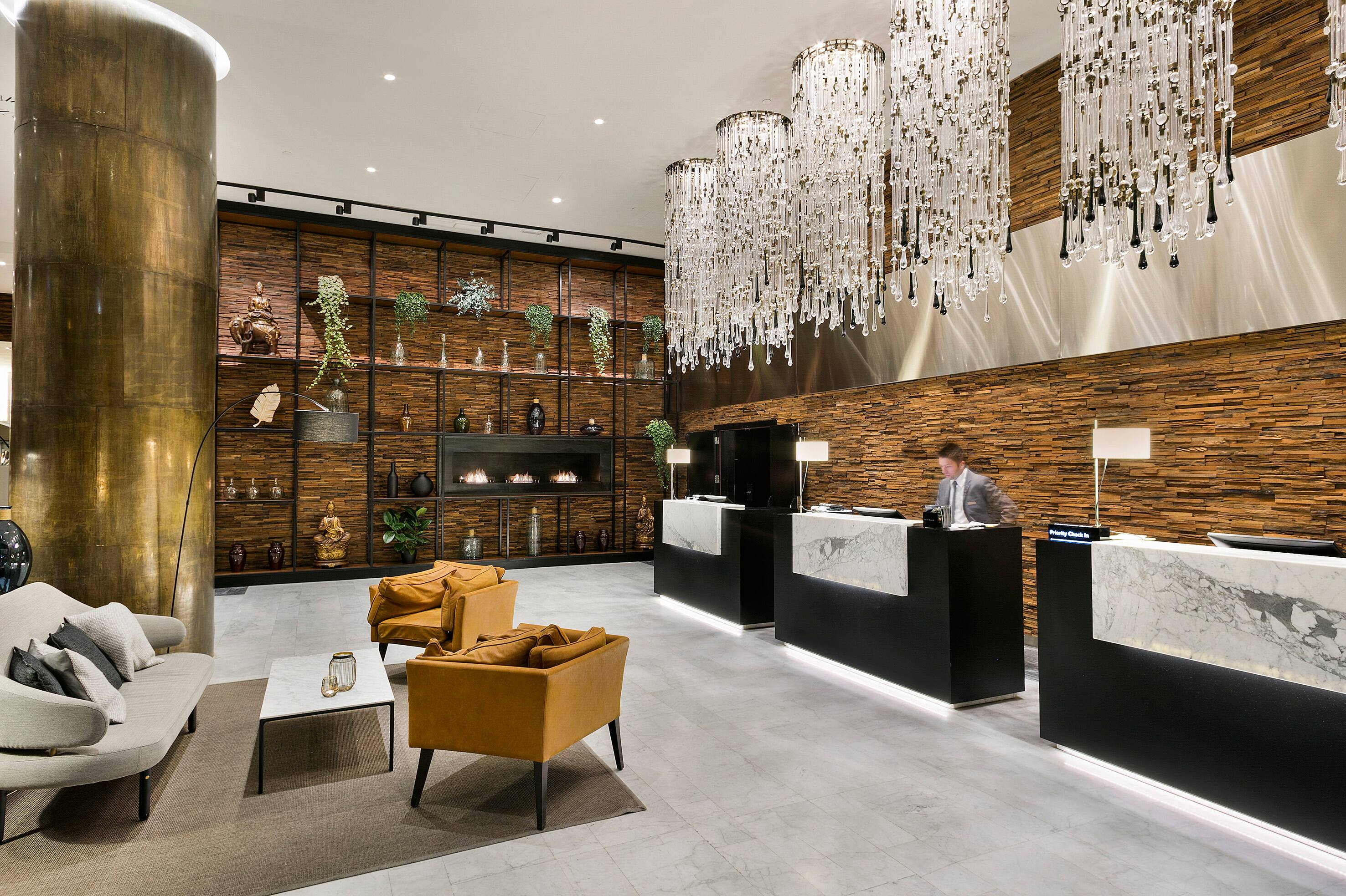 A hotel lobby with marble-fronted reception desks, cascading crystal chandeliers, a wood-slat wall and mustard armchairs around a low table.