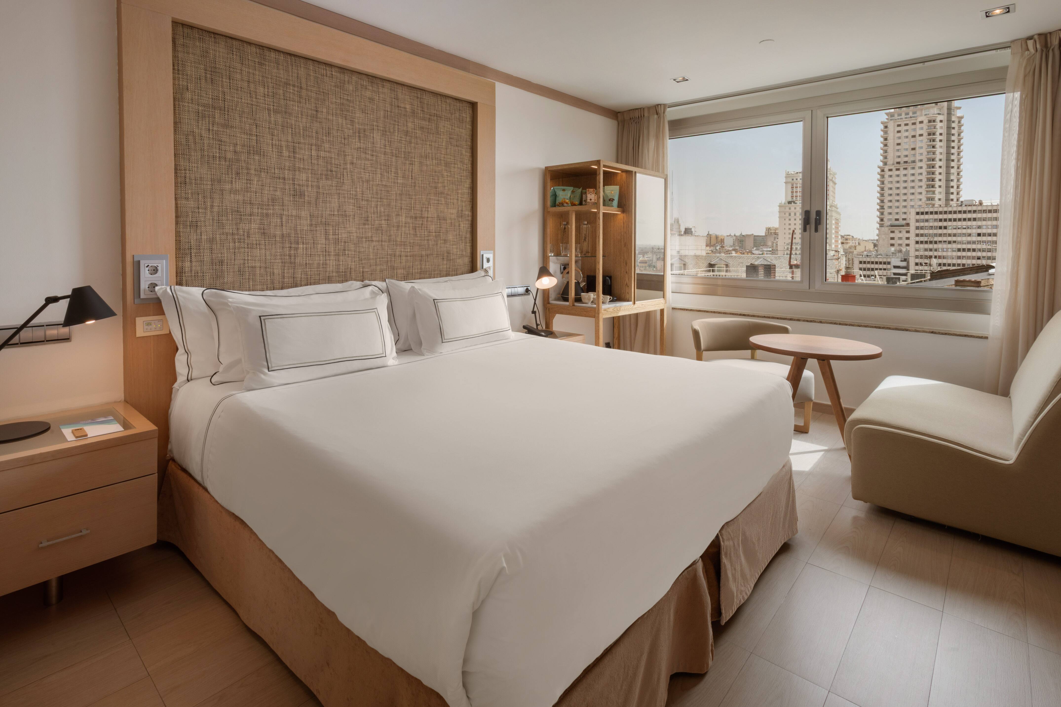 A guest room with a wide bed in white linens against a woven headboard panel, an armchair and a window looking out over high-rise towers.