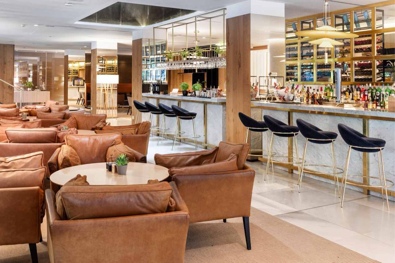 Marble bar counter backed by a lit wine wall, with dark velvet bar stools and tan leather lounge chairs in the foreground.