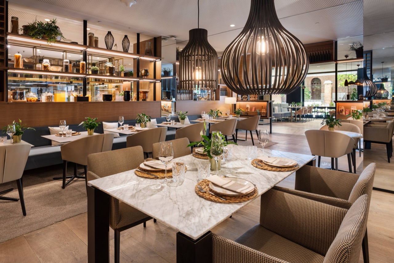 Arado Grocery & Restaurant dining room with marble-topped tables, woven placemats, large drum pendant lights, and an open pantry shelf in the background.