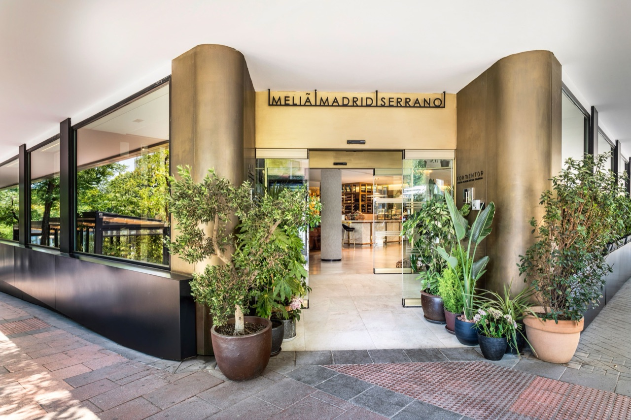 Ground-floor entrance of Meliá Madrid Serrano with bronze-clad columns, the hotel's sign, and potted olive trees flanking the glass doors.