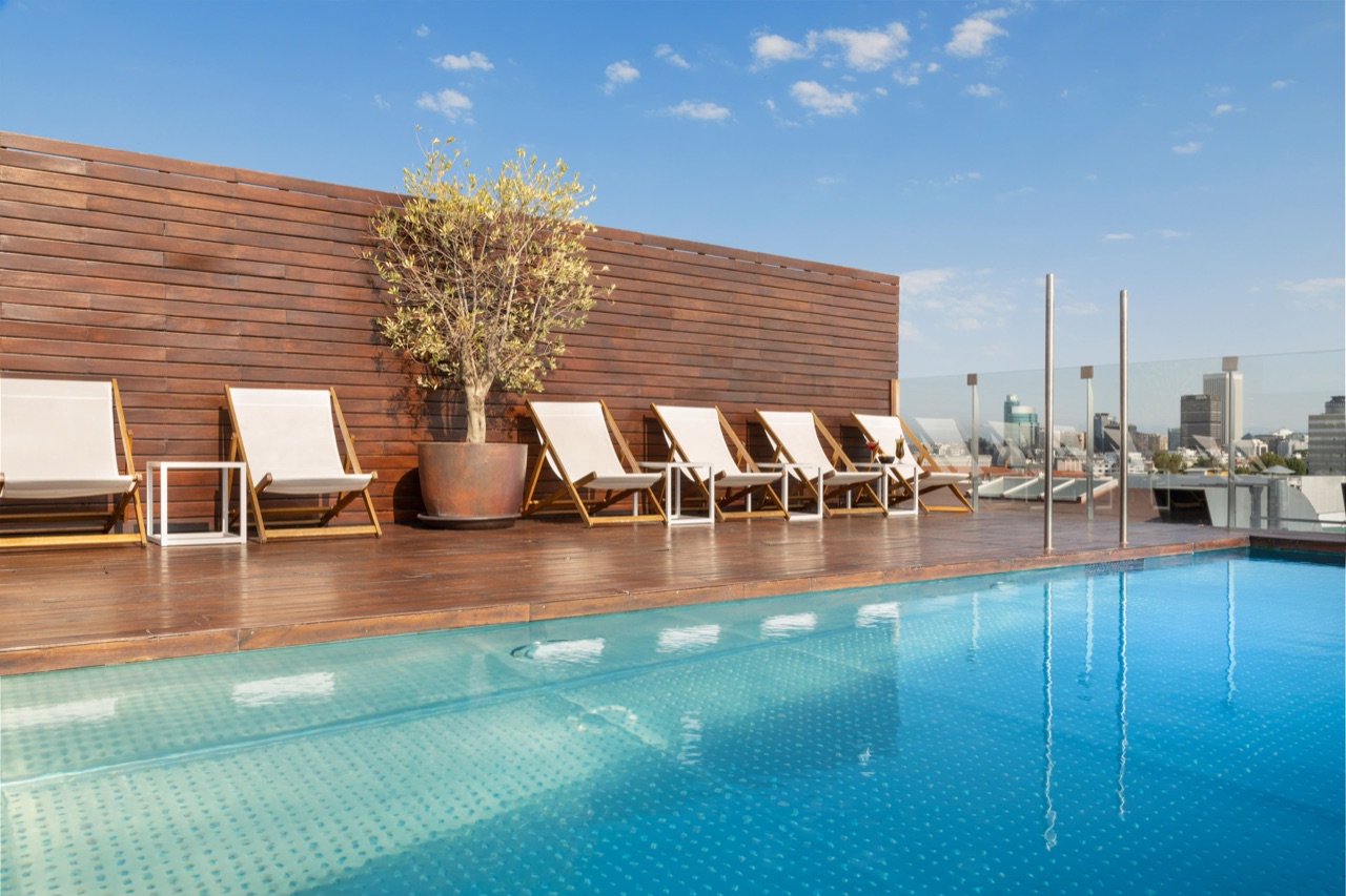 Rooftop pool with a wood deck, white sling-back sun loungers, and the Madrid skyline in the background under a clear sky.