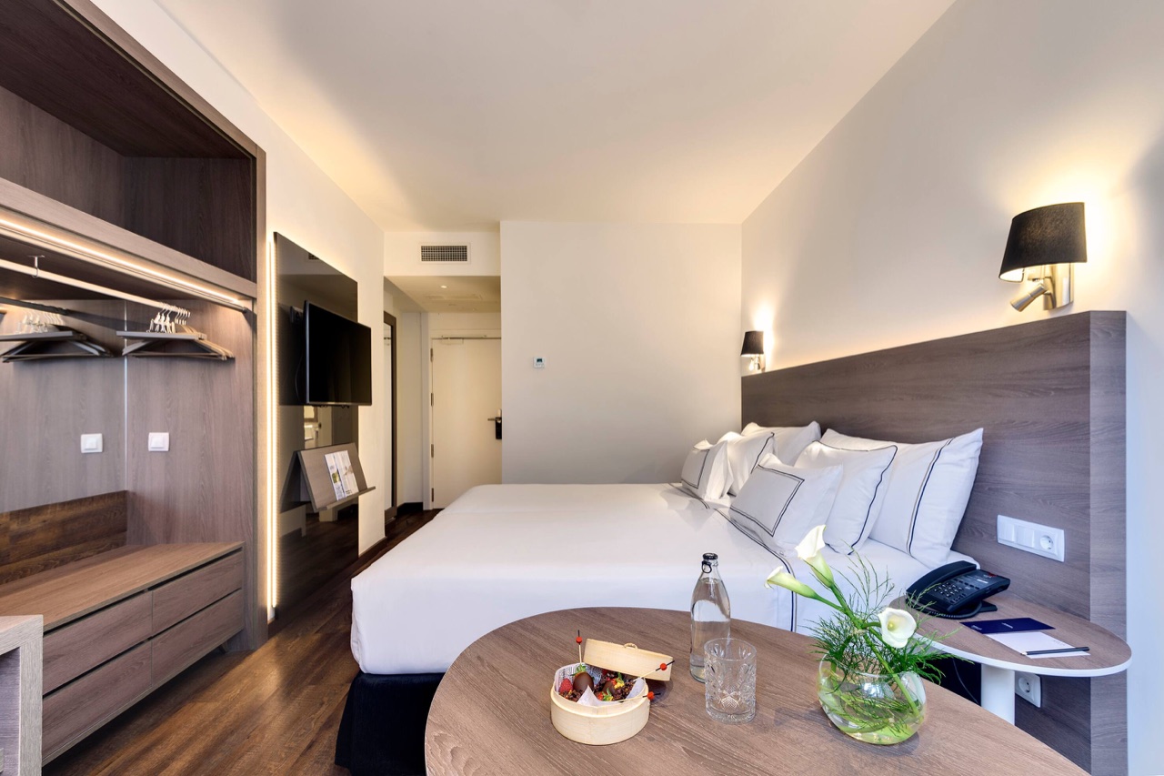 Hotel room with a white-linened double bed, a dark wood headboard, a wall-mounted TV, and a round table with a water bottle and fresh flowers.