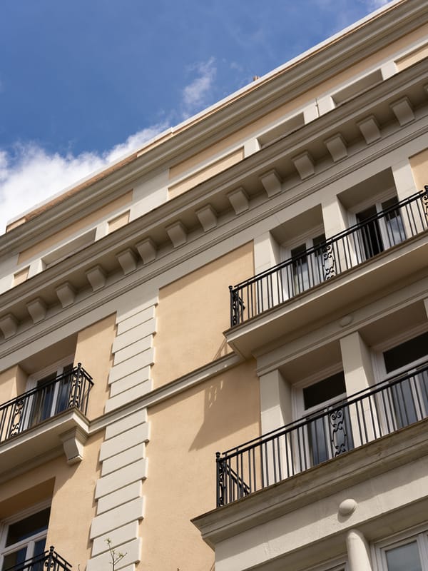 The hotel's cream stone facade seen from below, with black wrought-iron balconies, moulded window surrounds and a dentilled cornice against a blue sky.