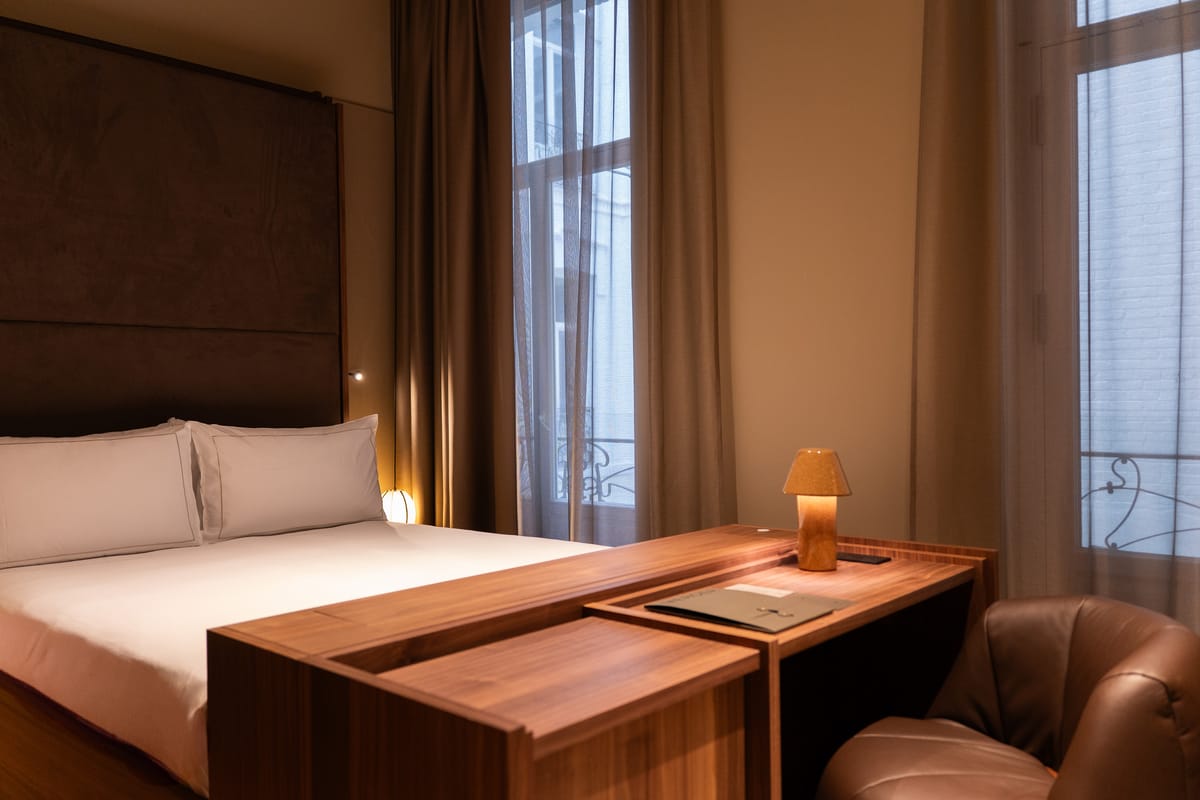 A double bed with white linens beside a walnut desk with a small lamp, tall windows opening onto wrought-iron balconies.