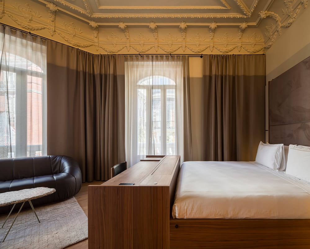 A corner room with an ornate plasterwork ceiling, arched windows behind sheer curtains, a suede headboard and a black leather sofa.