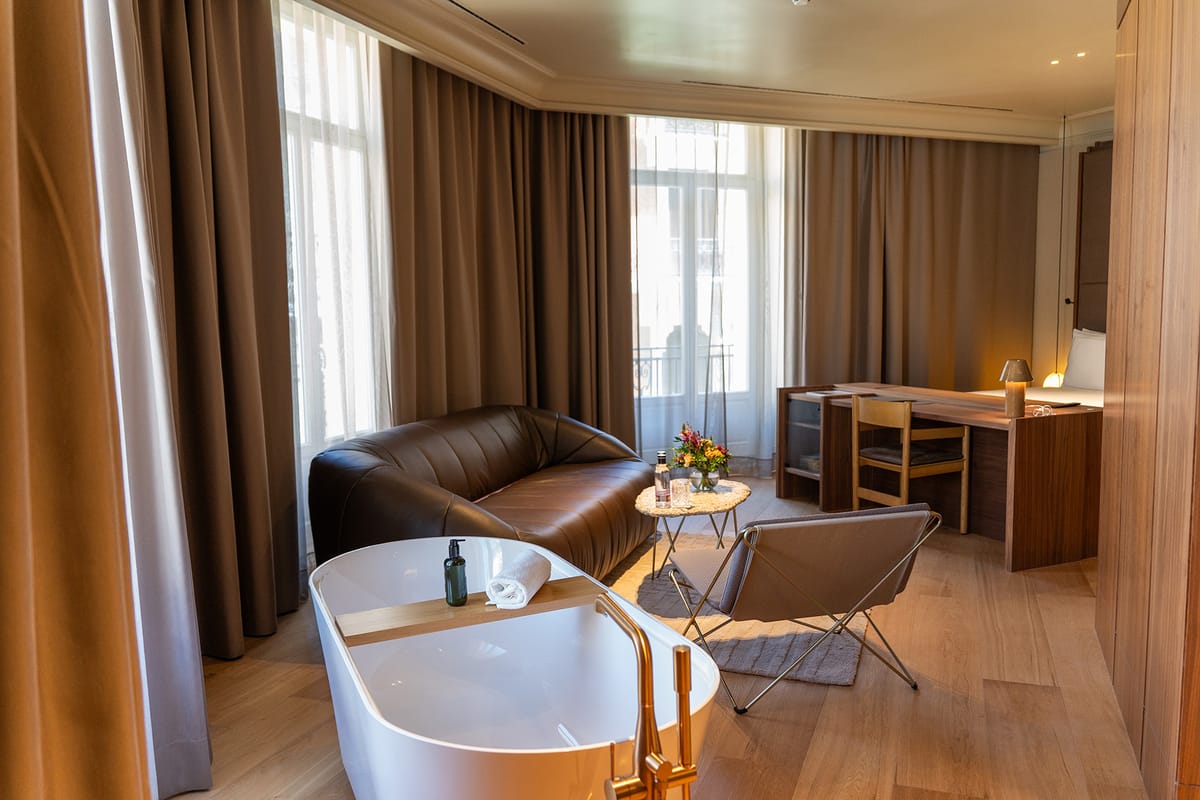 A freestanding white bathtub with brass taps beside a sitting area with a brown leather sofa, walnut desk and tall curtained windows.