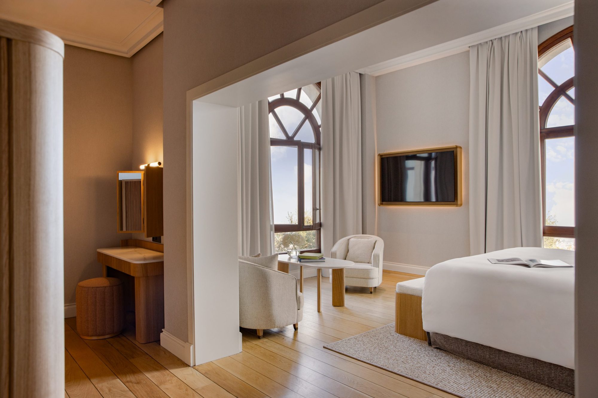 METT Suite bedroom at METT Barcelona with a white bed, wall-mounted TV, and a seating nook beneath a tall arched window