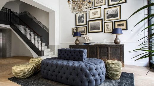 Lobby lounge at Midmost Barcelona with a tufted navy velvet daybed, gold bouclé poufs, a gallery wall of framed prints, and a black iron staircase