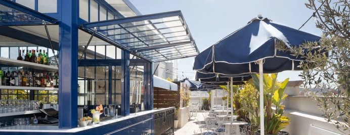 La Fresca rooftop bar at Midmost Barcelona with a backlit glass-walled bottle display and navy sun umbrellas over the terrace seating