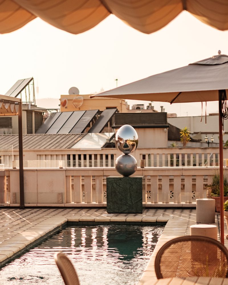 Rooftop pool at Borneta with a striped umbrella and a metallic sphere sculpture beside the water, Barcelona rooftops visible beyond