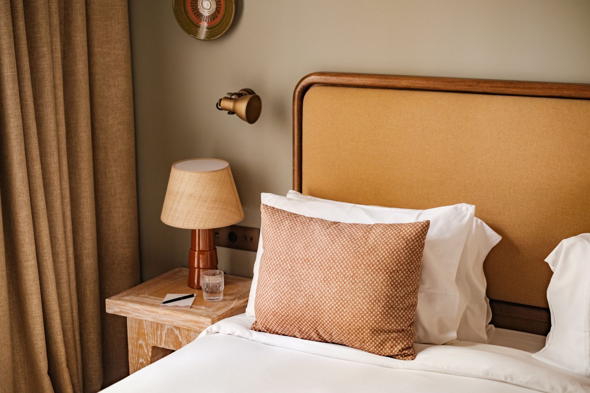 Guest room detail at Borneta with a curved walnut-toned wood headboard, a terracotta bolster cushion and a wicker table lamp on a wood nightstand