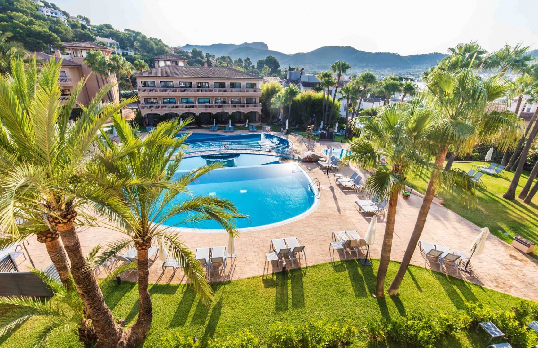 Aerial view of the hotel's curved outdoor pool surrounded by palm trees and sun loungers, with the Tramuntana mountains behind