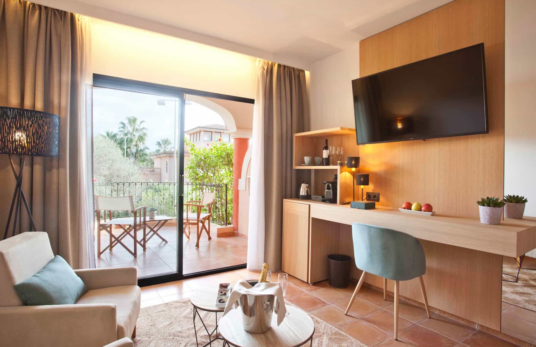 Junior suite living area with a wall-mounted TV, wood shelving unit and a private terrace overlooking the gardens
