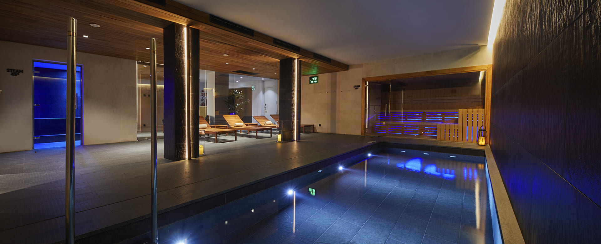 Indoor spa pool at night with blue mood lighting, wooden loungers and a glass-walled sauna