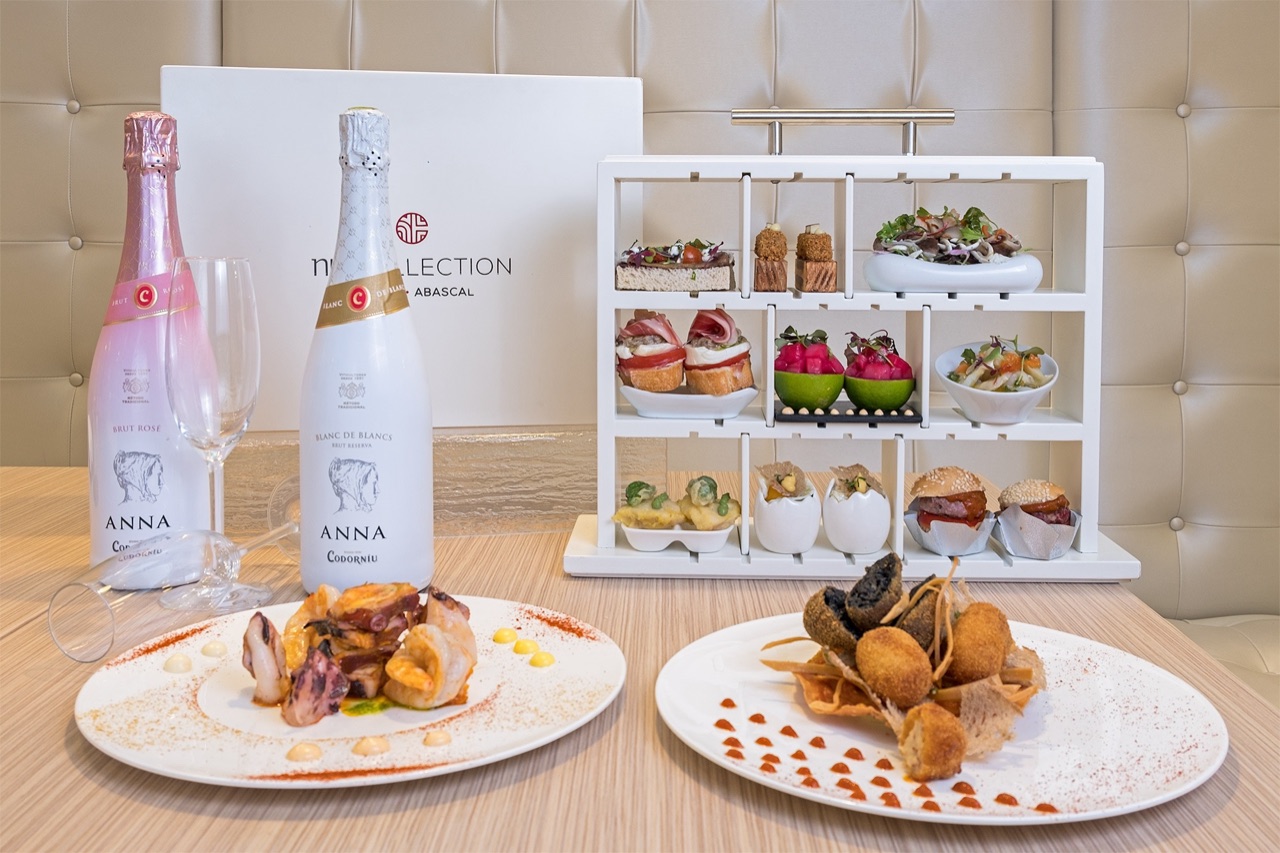 DOMO's The Box tasting stand with small tapas plates arranged on a tiered white rack, next to two plates of seafood and a branded NH Collection Madrid Abascal box on a booth table.