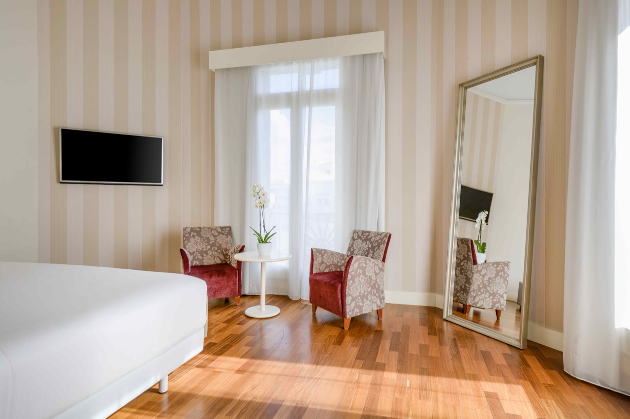 Hotel room with striped cream wallpaper, two floral-pattern armchairs around a small round table by a curtained balcony door, a wall-mounted TV and a full-length mirror.