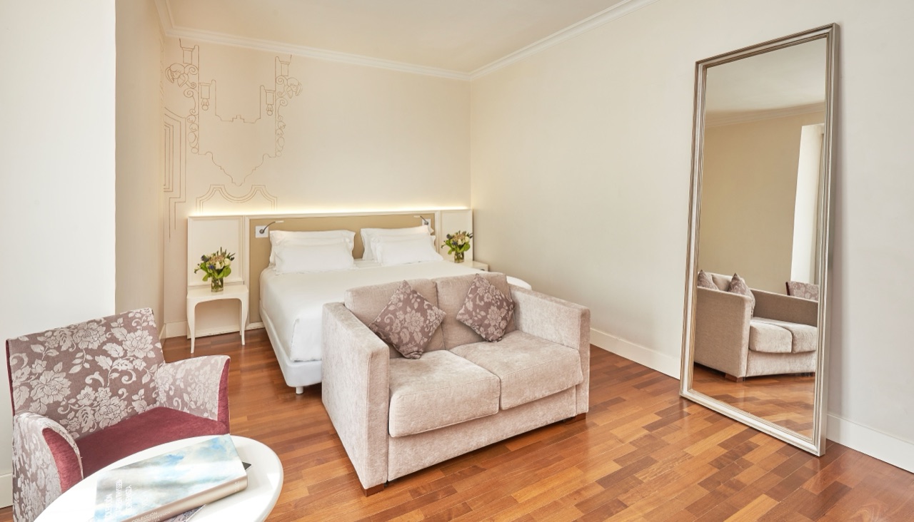 Hotel room with a line-drawing mural of a Madrid building above the bed, a beige loveseat at the foot of the bed and a large floor mirror against the wall.