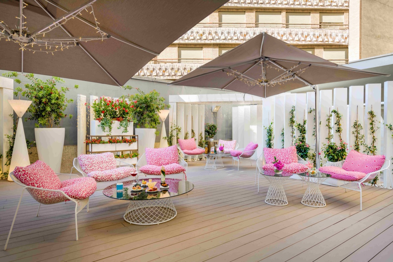 Outdoor terrace lounge with wire-frame armchairs in pink patterned cushions, glass coffee tables, large canvas umbrellas and potted plants against a white fence.