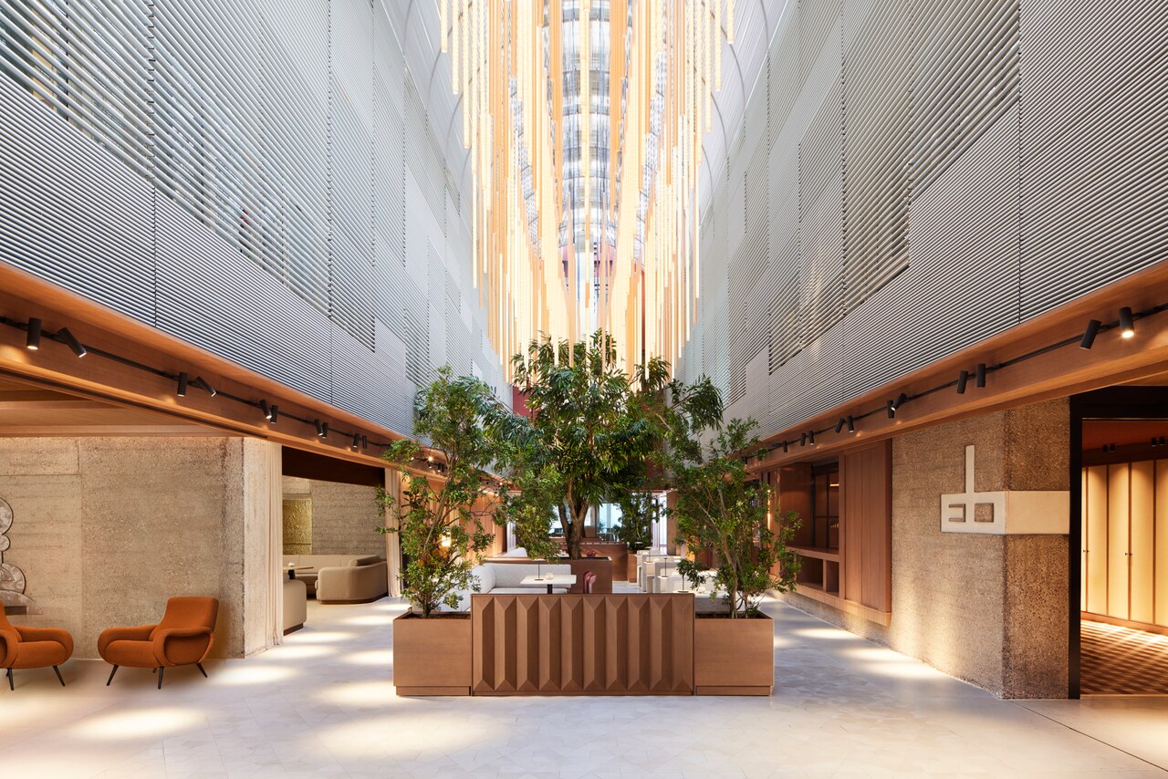 A soaring hotel lobby atrium with concrete walls, a hanging light installation and trees planted in wooden planters at the center.