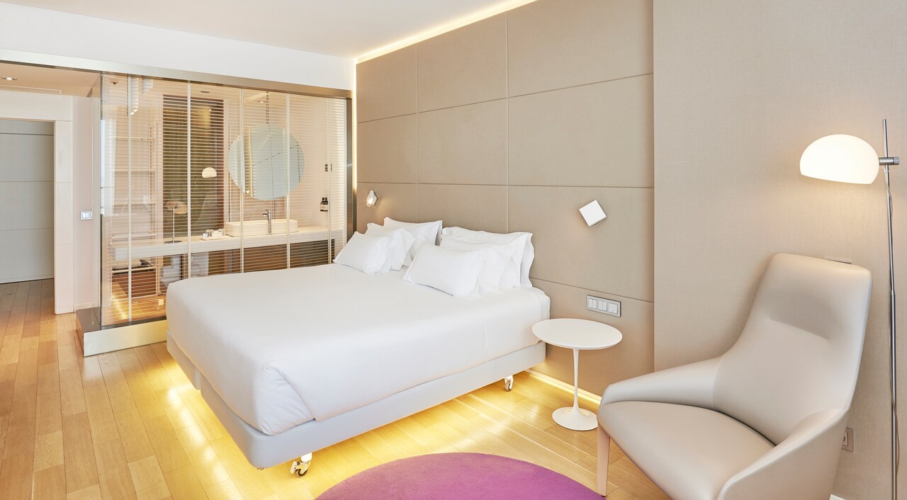 A hotel room with a white upholstered bed, a glass-walled bathroom with a round mirror, and a beige leather armchair.