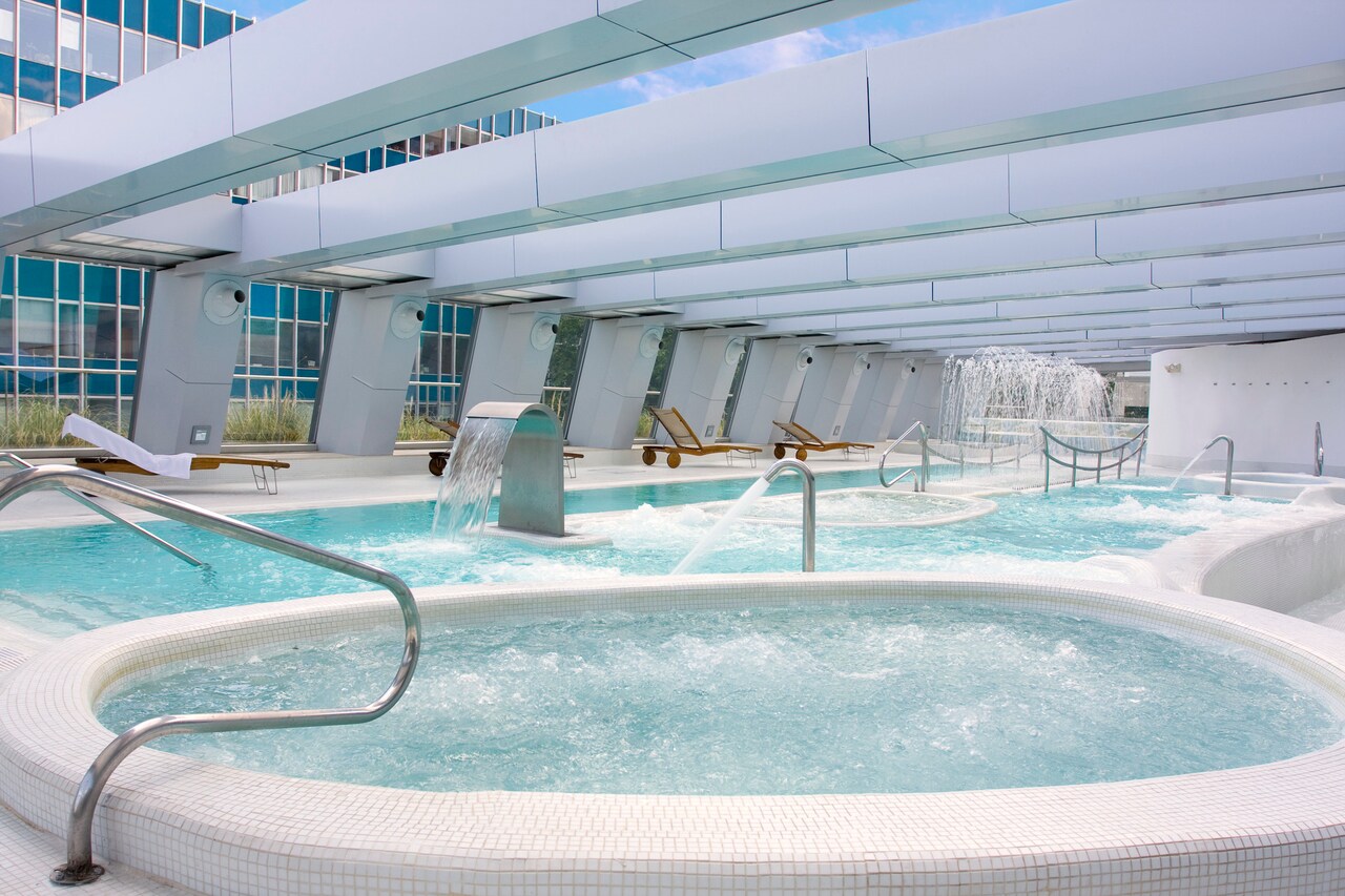 An outdoor spa terrace with hydromassage pools, jet fountains and loungers, overlooked by the hotel's glass tower.