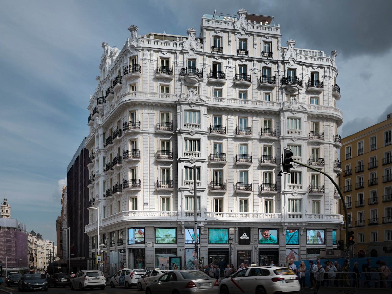 The hotel's cream and white corner building with wrought-iron balconies on every floor, shop windows at street level and taxis passing.