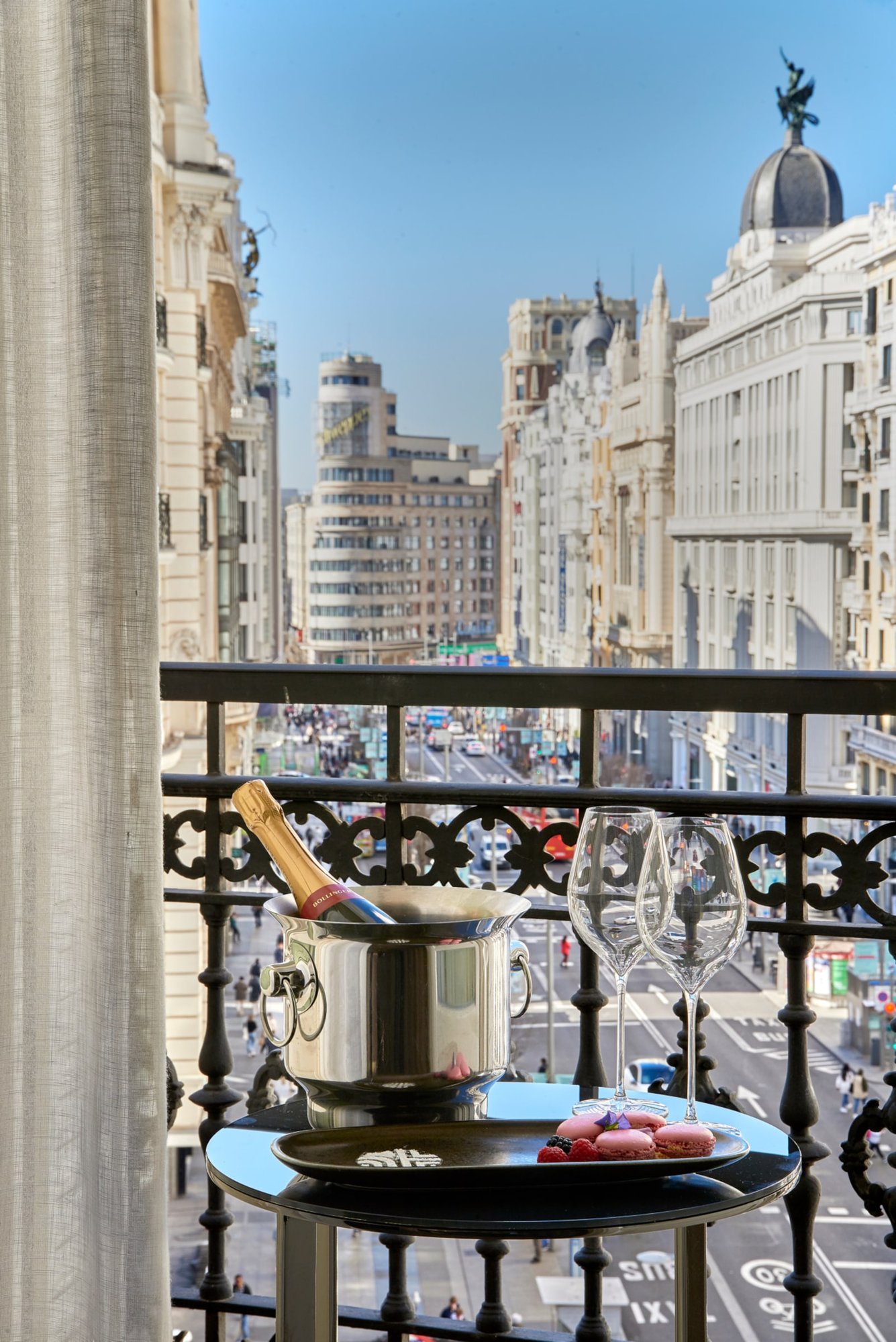 A wrought-iron balcony with a champagne bucket, two glasses and macarons on a small table, looking down a long avenue of white stone buildings.