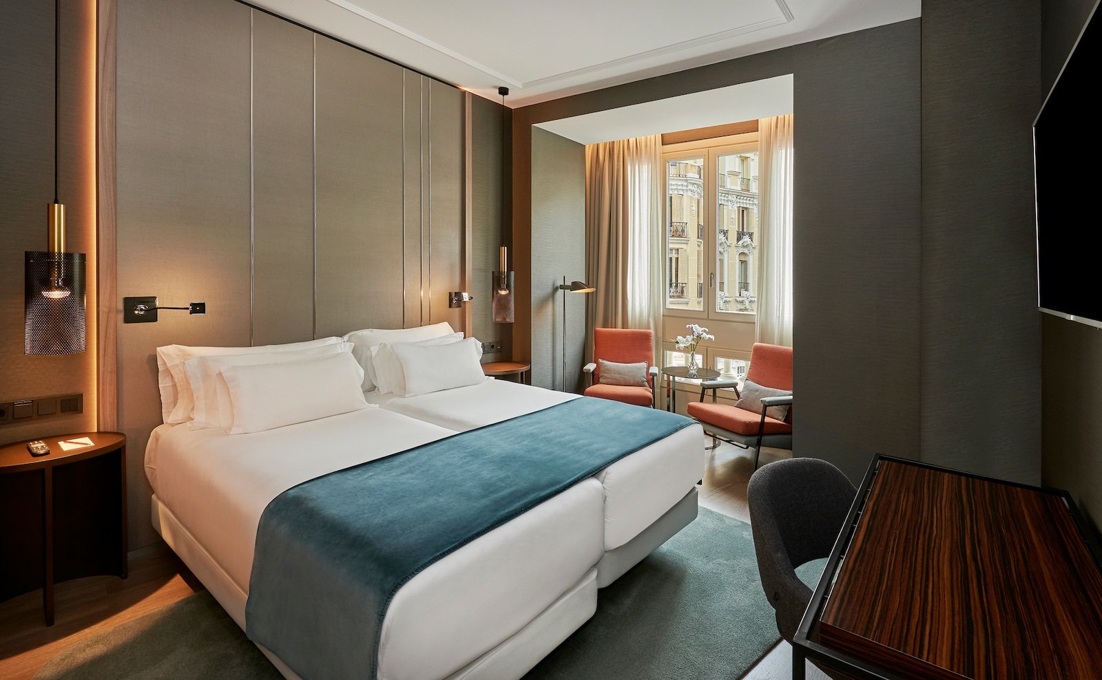 A twin-bed room with a teal bed runner, a taupe panelled headboard and two orange armchairs beside a tall window onto neighbouring white stone buildings.