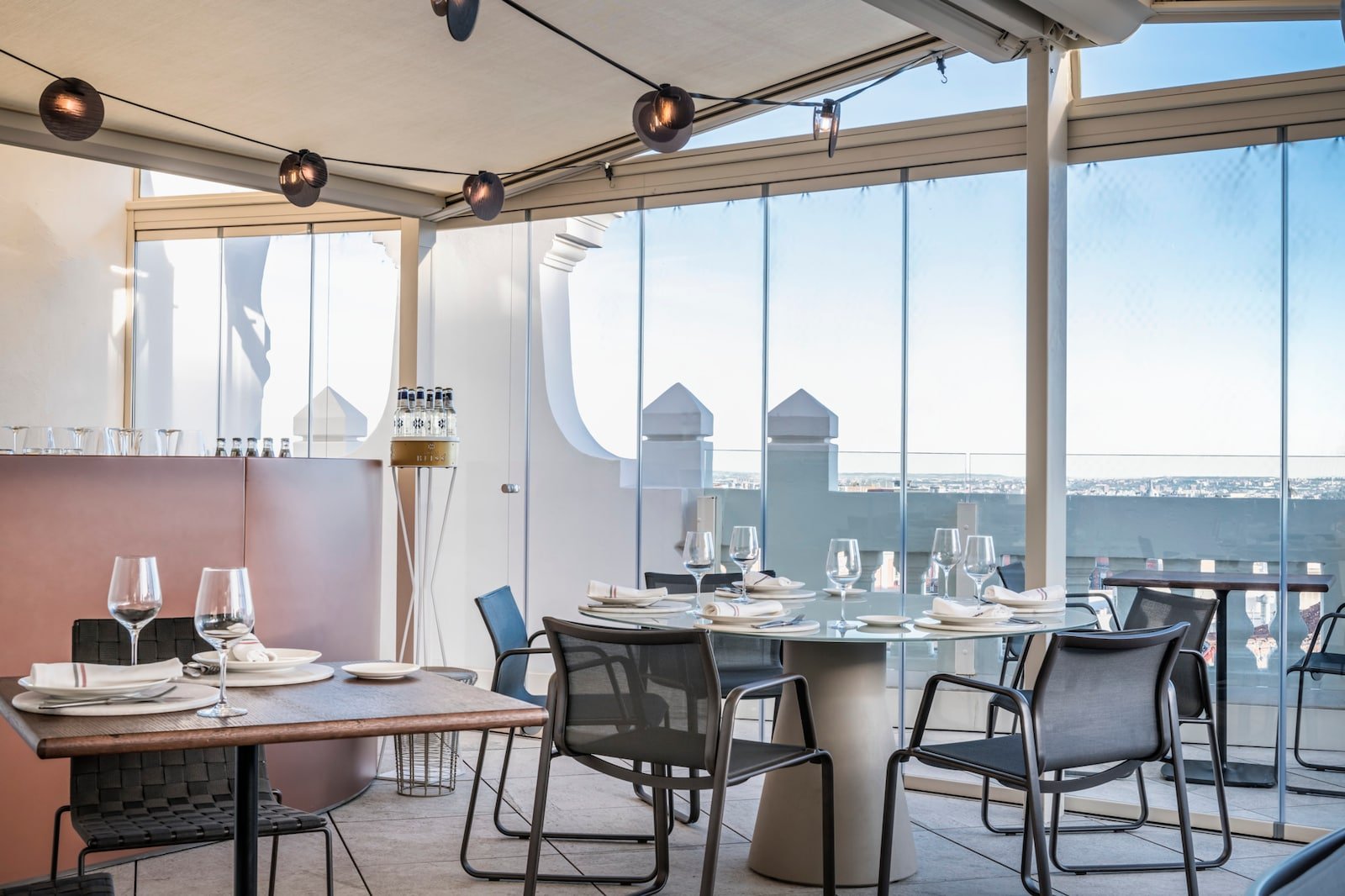 A glass-enclosed terrace dining area with tables laid for service and a wide view of the city beyond.