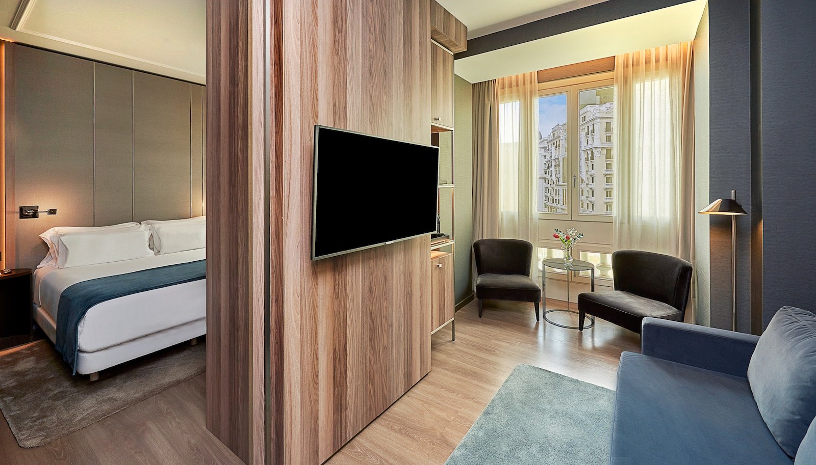 A suite-style room with a wooden partition holding a wall-mounted TV, a bed on one side and blue armchairs and a sofa by a window on the other.