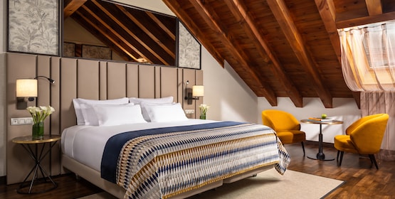 An attic guest room with exposed wooden roof beams, a padded headboard wall, a patterned bedspread and two mustard armchairs at a small table.
