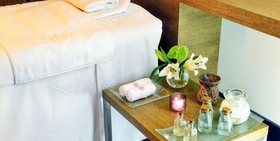 A spa treatment table with folded towels beside a side table holding rolled towels, small bottles of oil, a candle and flowers.