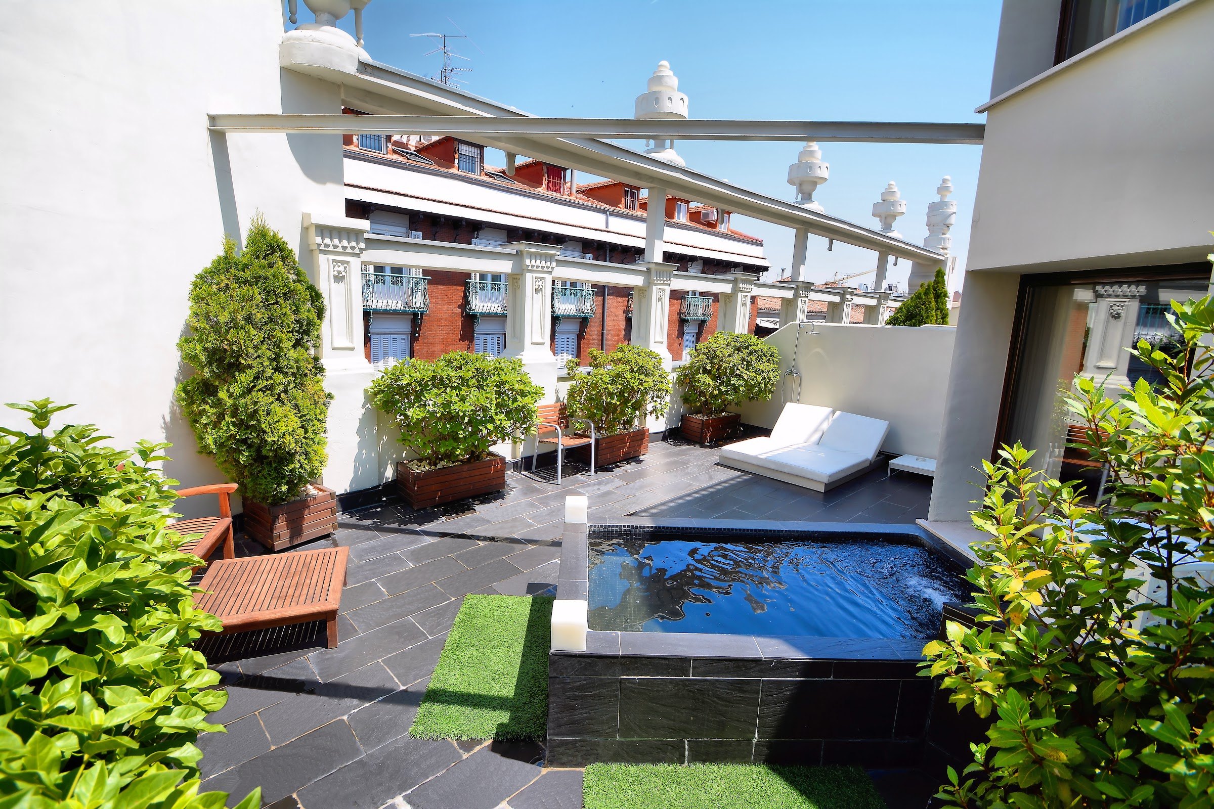 A roof terrace with a small plunge pool, potted shrubs, a white daybed and wooden loungers between white walls.