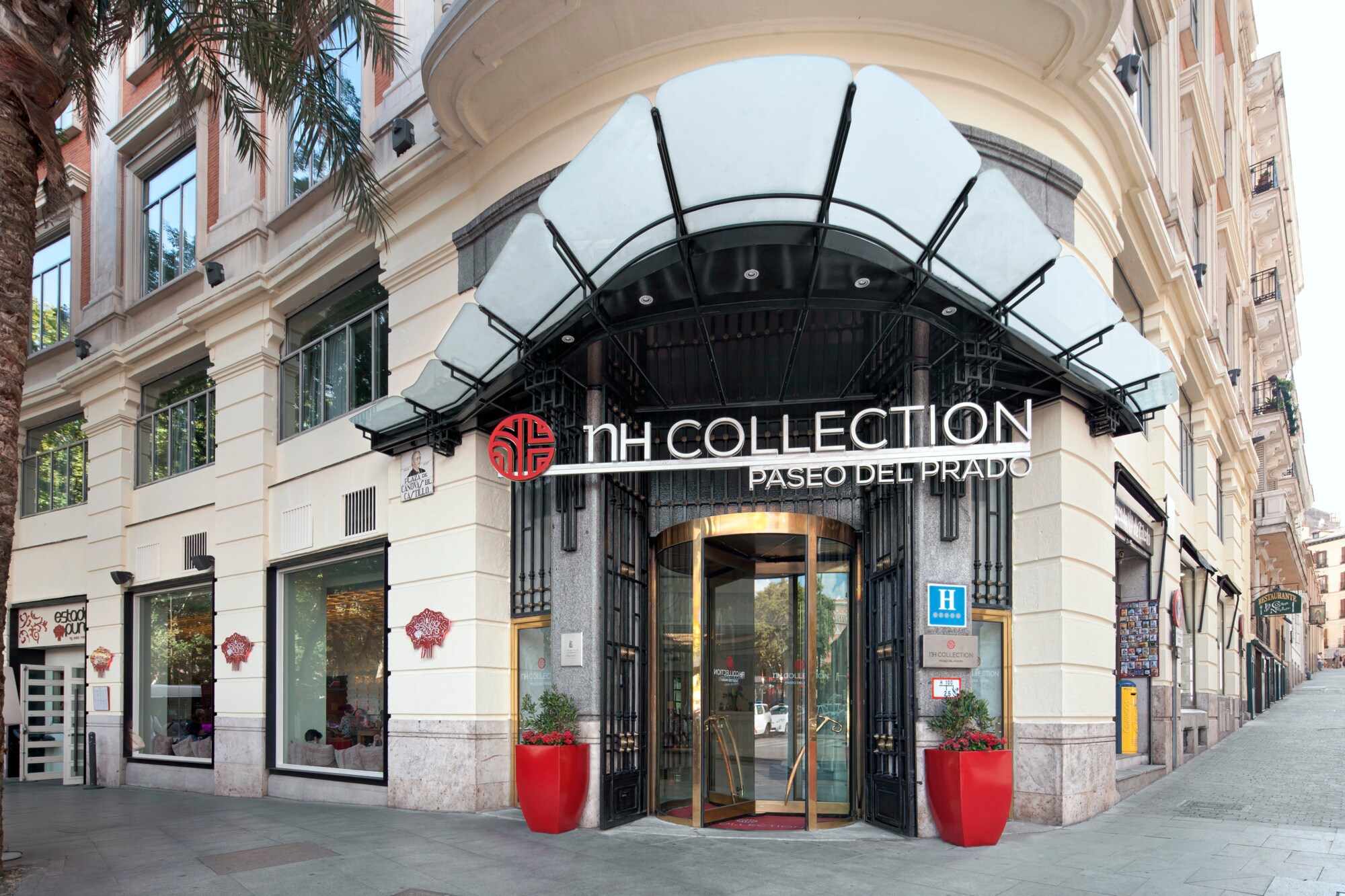 The hotel's corner entrance beneath a fan-shaped glass-and-iron canopy, with NH Collection Paseo del Prado lettering above a brass revolving door flanked by red planters.