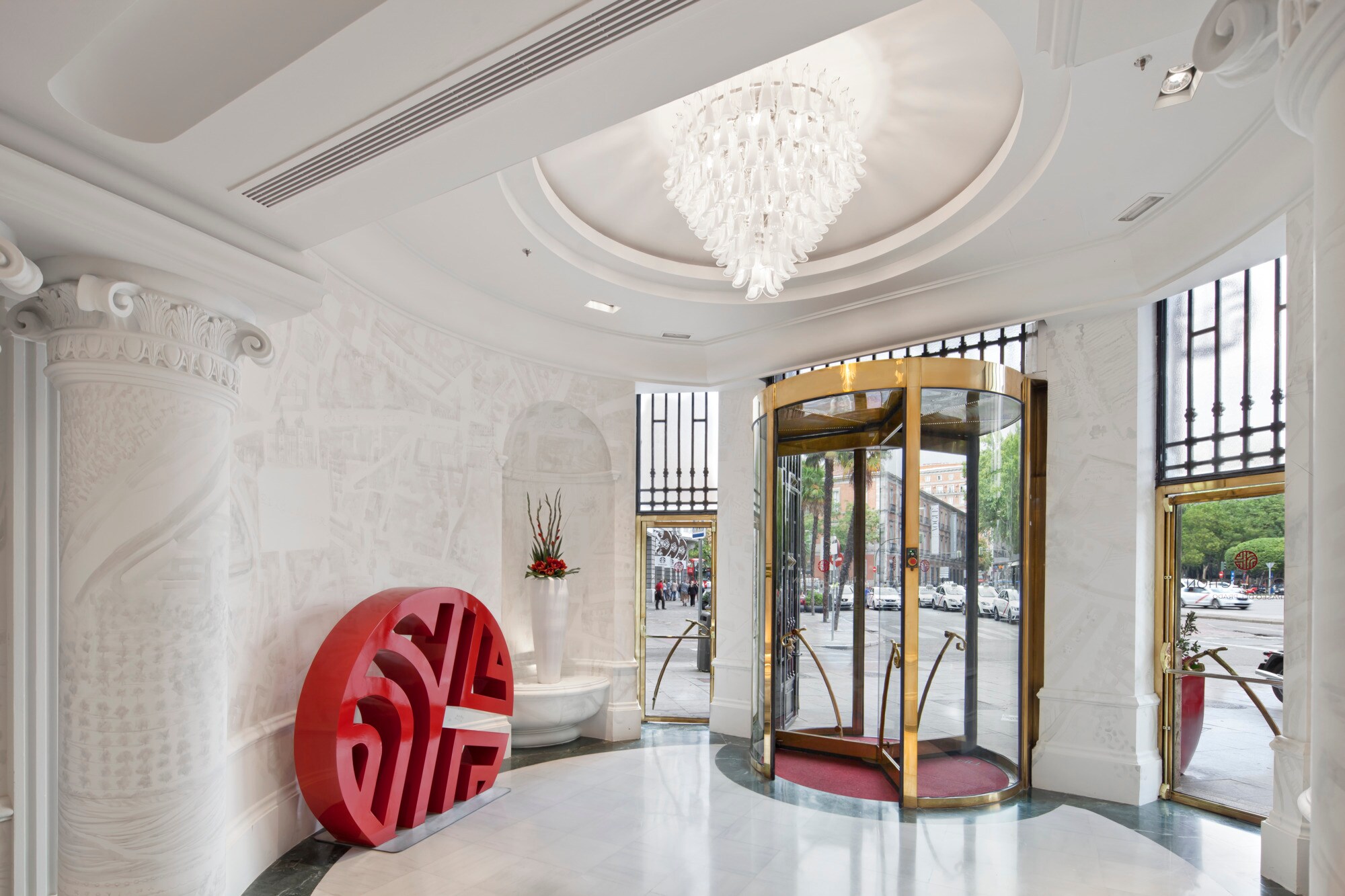 The entrance rotunda with white relief-carved walls and columns, a glass-petal chandelier in a domed ceiling, a brass revolving door and a red NH monogram sculpture.