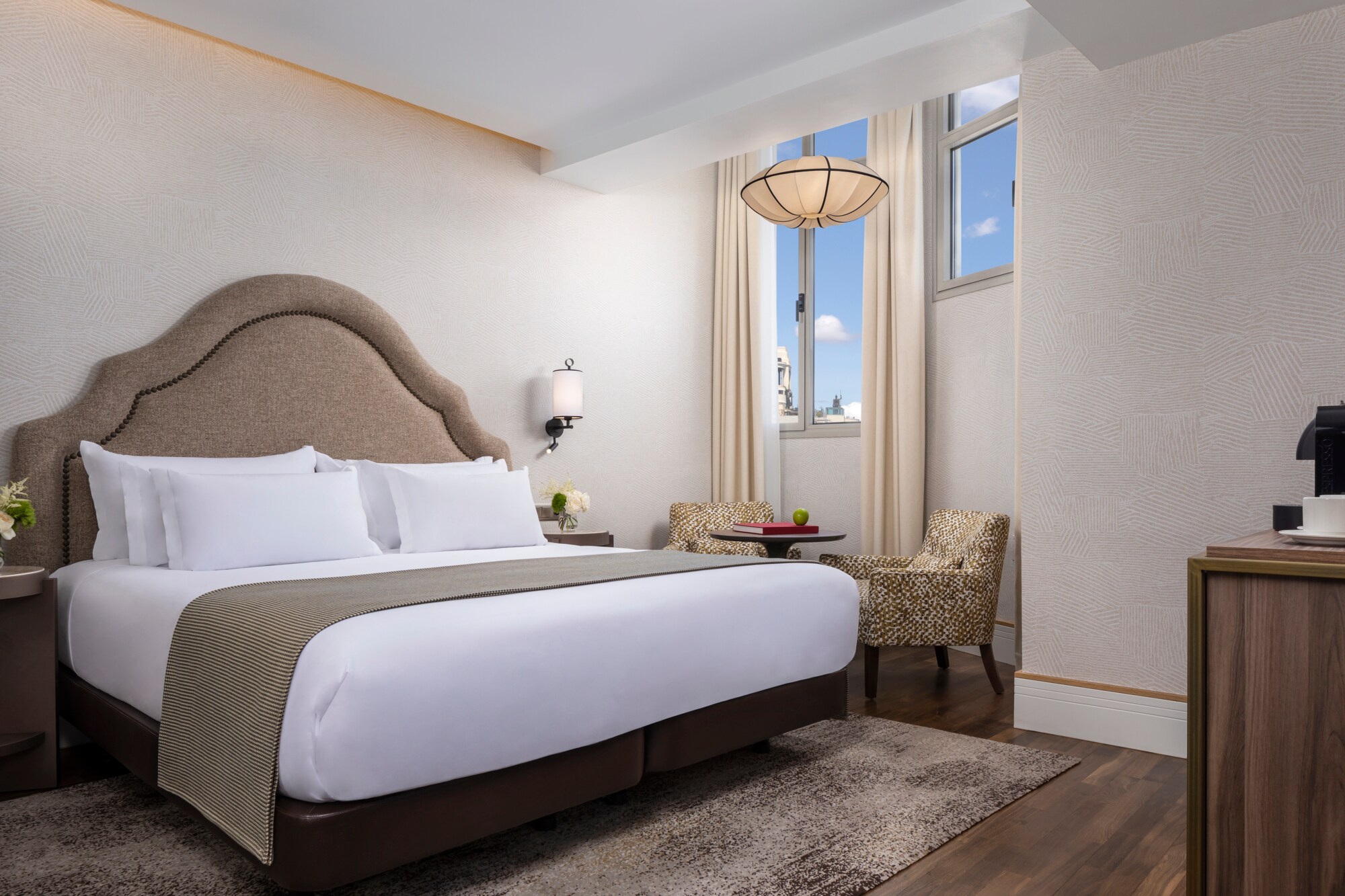 A guest room with an arched studded-fabric headboard, white bedding with a taupe runner, two patterned armchairs around a small table and a window with a city view.