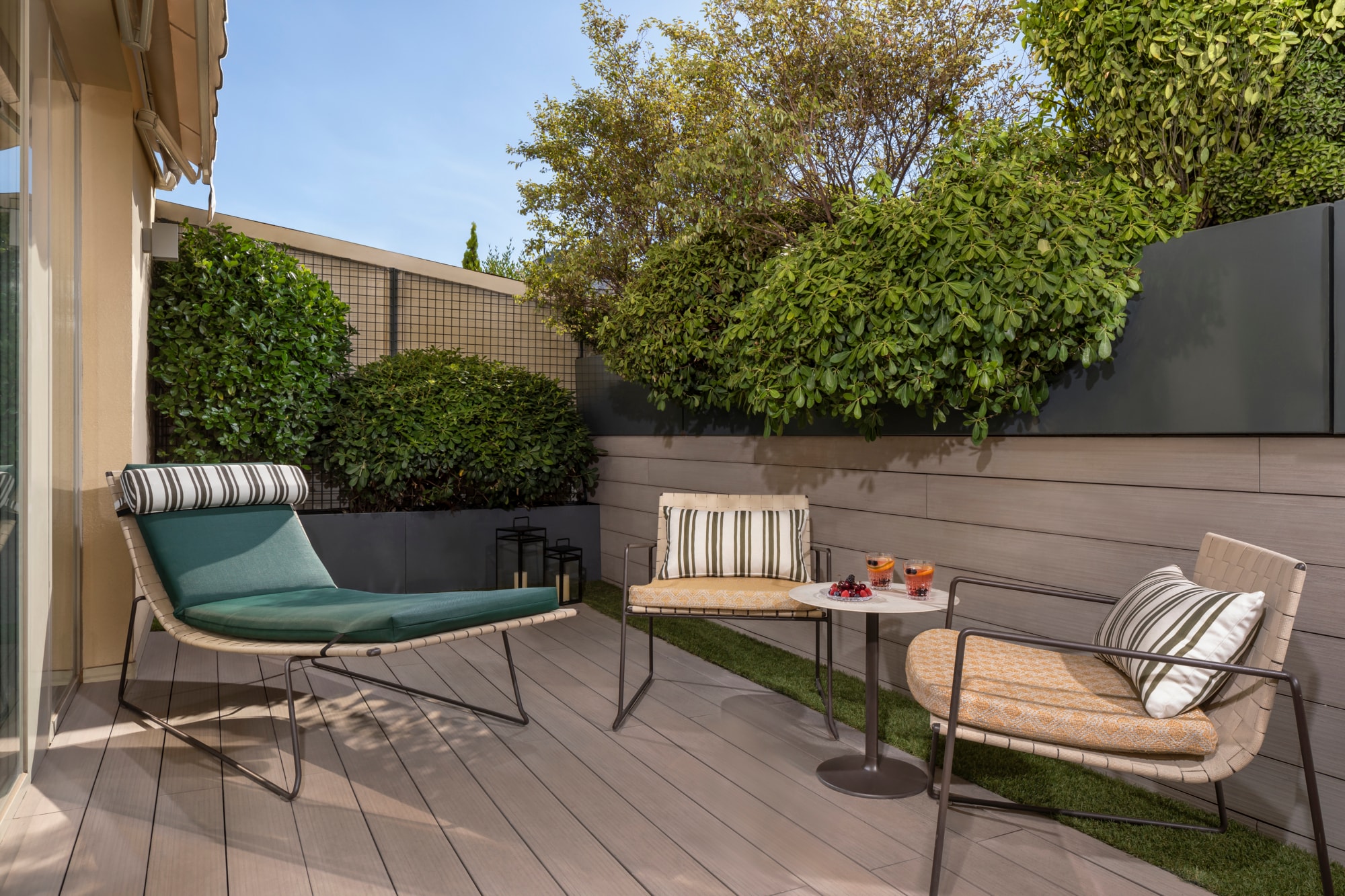 A room terrace with a green-cushioned lounger and two woven armchairs around a small marble table holding two drinks and a bowl of berries, on wooden decking enclosed by planted greenery.