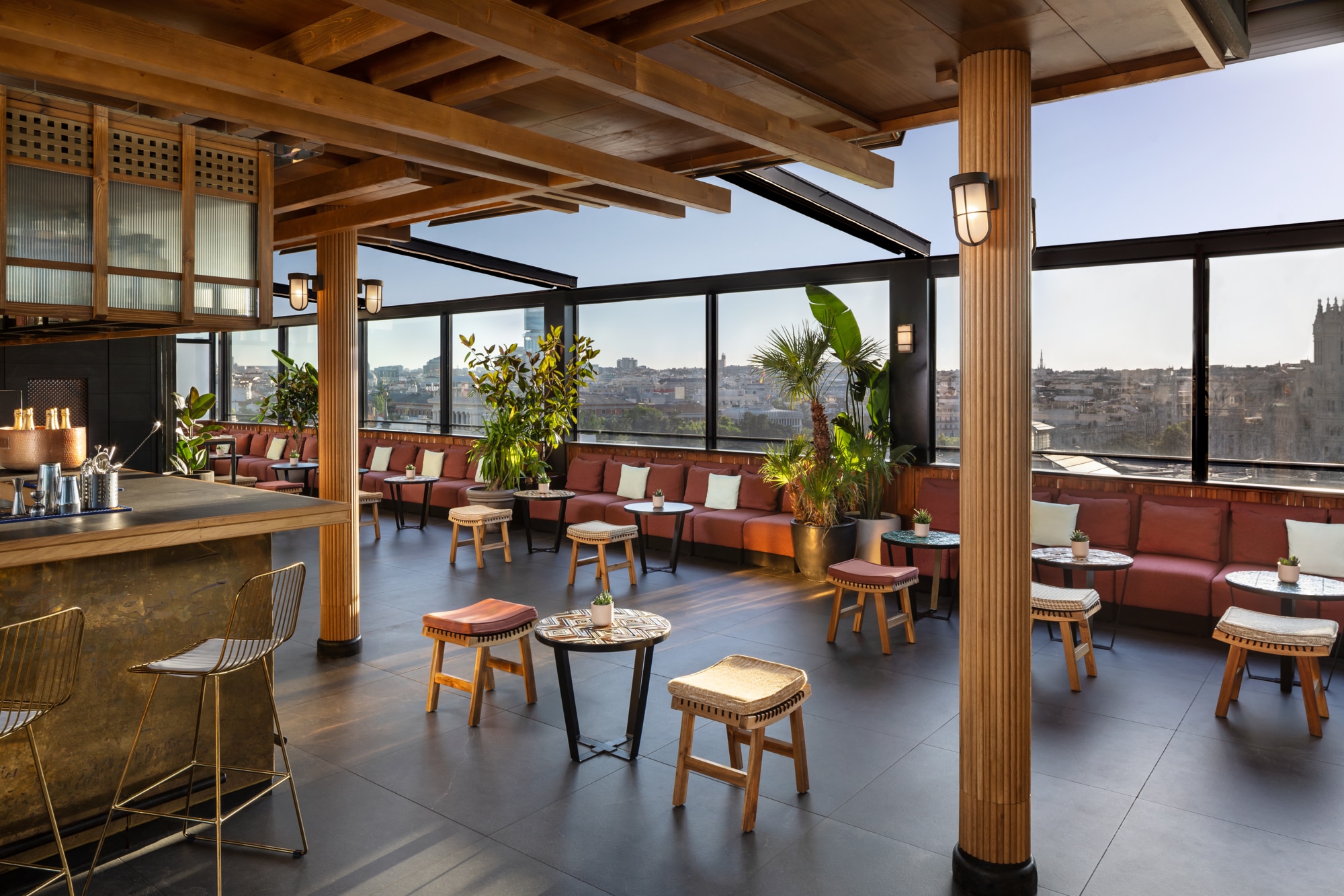 The covered rooftop bar with a brass-topped counter, wire barstools, wooden pillars, cushioned benches and stools, and glass walls looking over the Madrid skyline.