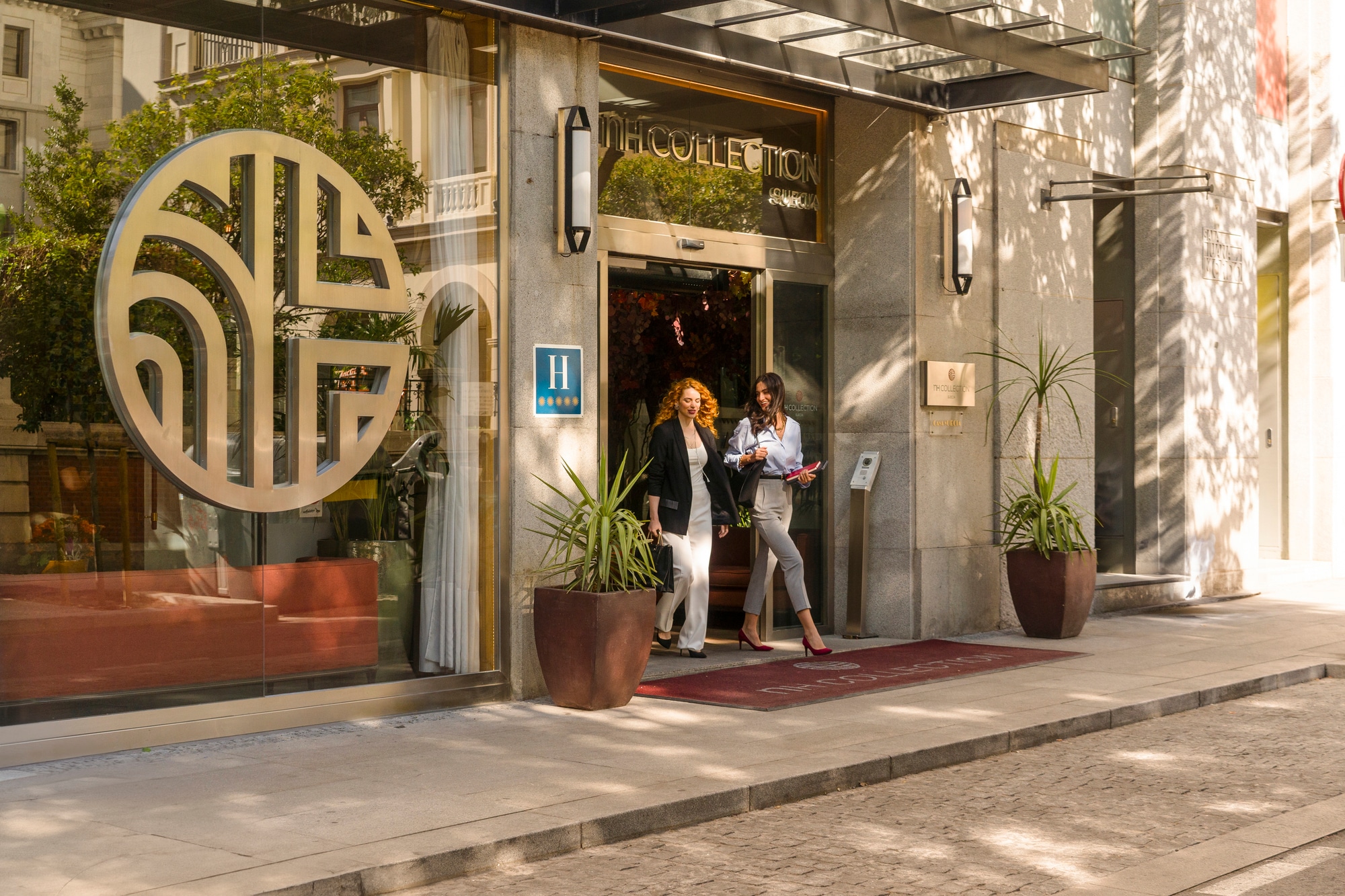 The hotel's street entrance with NH Collection Suecia lettering above the glass doors, a large circular brand monogram on the glazed facade and a red carpet at the doorway.